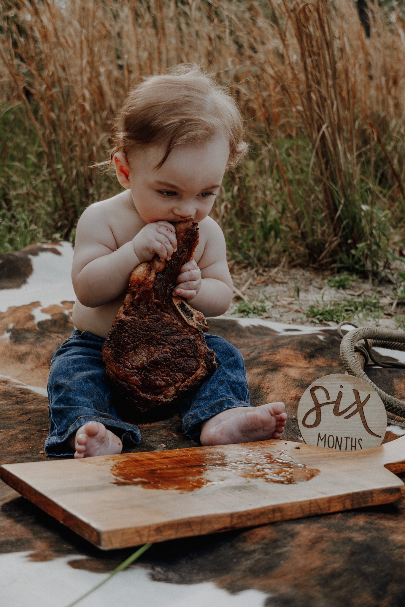 A young child with light brown hair, shirtless and wearing dark blue pants, sitting outdoors on a piece of animal hide, eating a large piece of cooked meat. In the background, there are tall, dry grasses, and on the ground near the child, there's a r