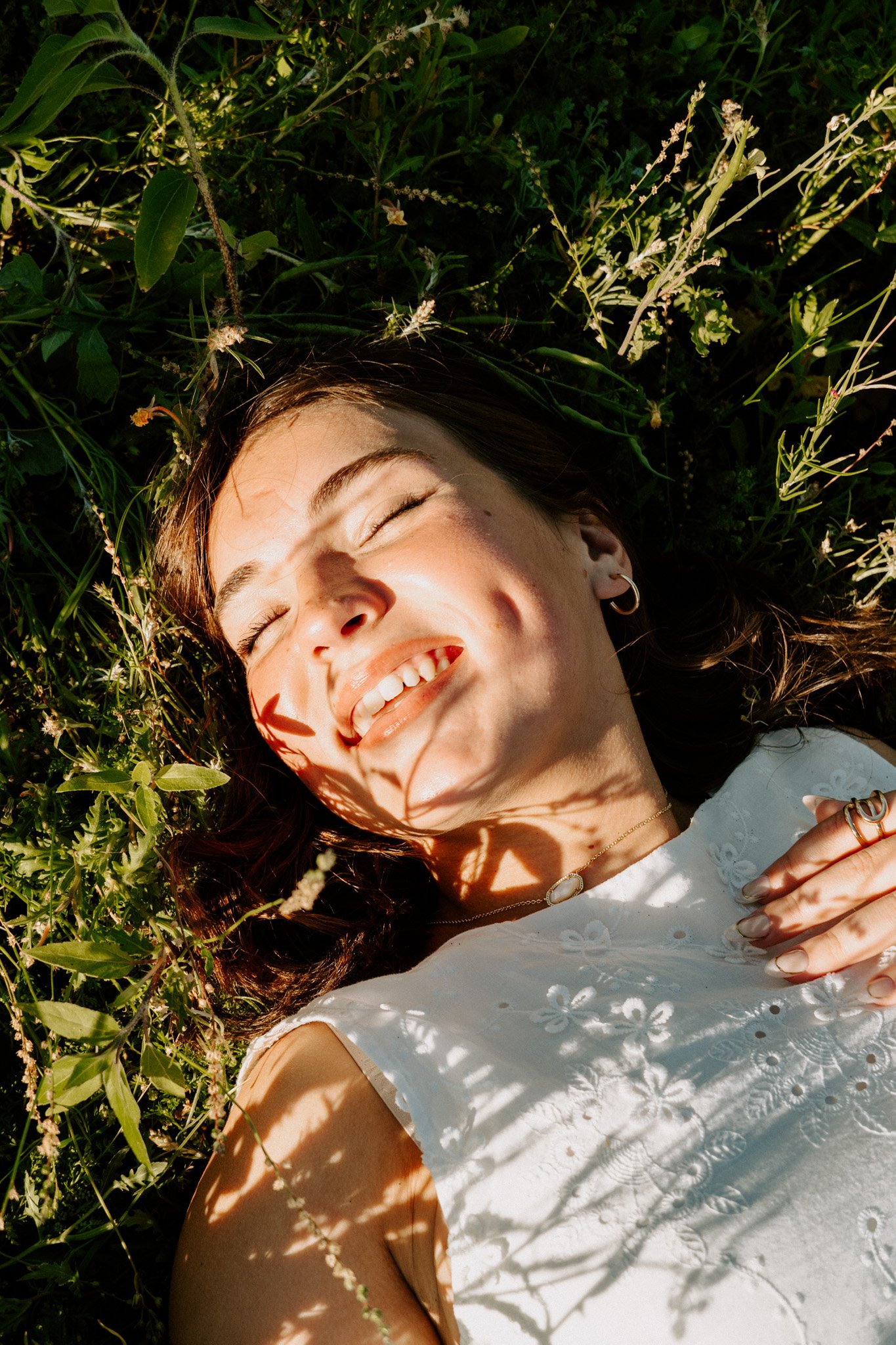A woman with light skin and brown hair lying on her back in a bed of green plants and grass, smiling with her eyes closed, wearing a white embroidered dress, jewelry, and receiving sunlight that creates shadow patterns on her face and dress.