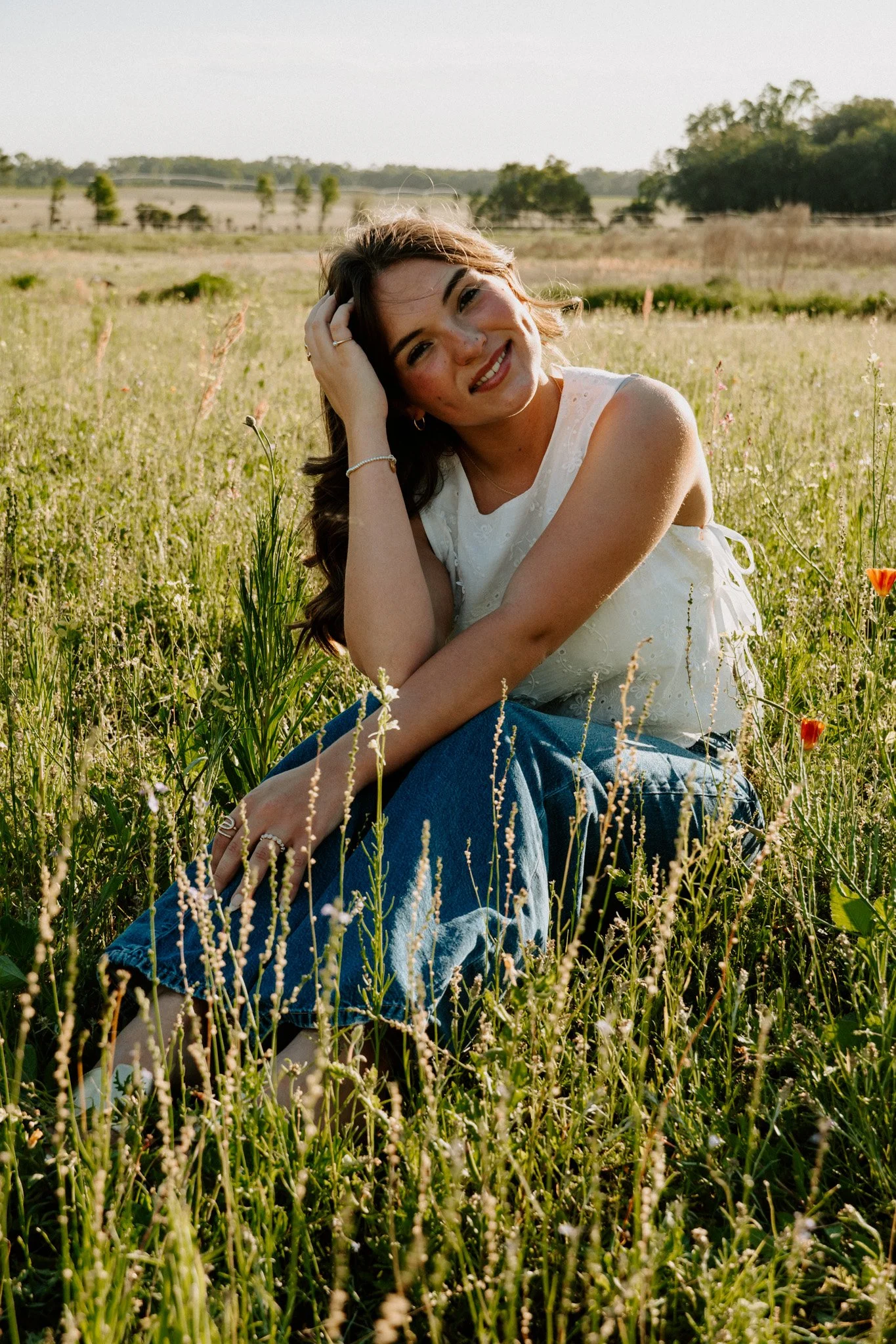 A young woman with long brown hair, wearing a white top and blue jeans, sitting in a grassy field with wildflowers, smiling and leaning her head on her hand, in a rural landscape with trees and a fence in the background during daytime.