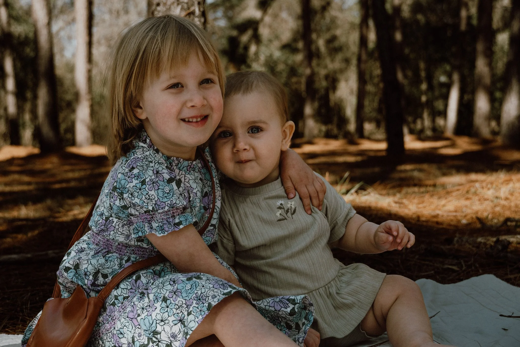 Two young children, a girl with red hair and a boy with light brown hair, sitting outdoors in a forest with trees and fallen leaves, smiling and hugging.