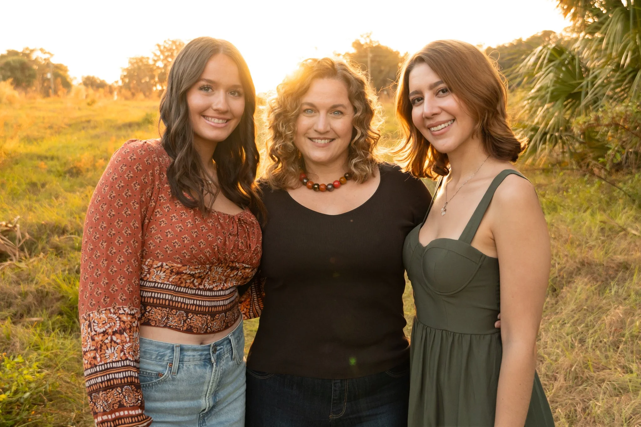 Three women standing outdoors during sunset, smiling, with trees and a grassy field in the background.