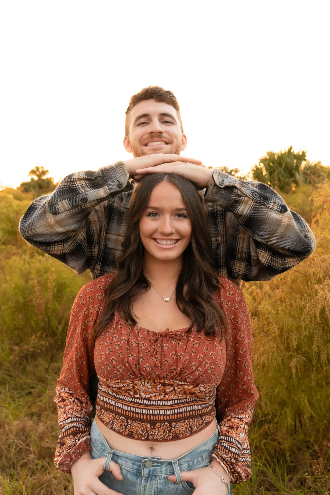 A young man and woman standing outdoors in a field during sunset, smiling at the camera. The man is behind the woman, resting his hands on her head, and the woman is wearing a patterned crop top, with her hands in her pockets.
