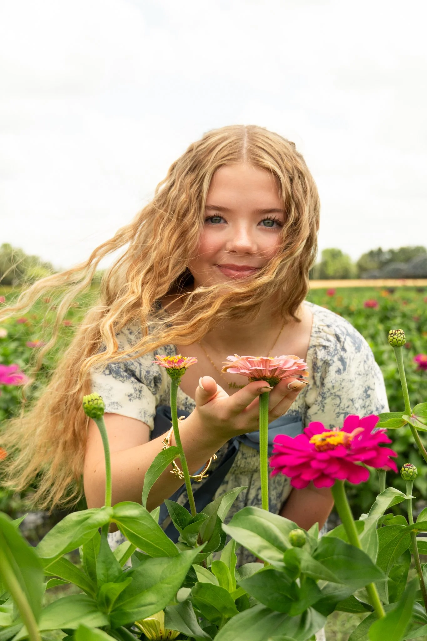A young woman with long curly hair holding a pink flower in a garden with various colorful flowers.