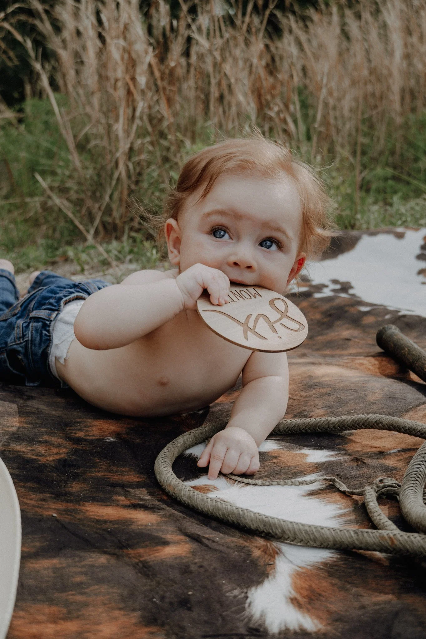 A young child with reddish hair and blue eyes lying on a wooden surface outdoors, holding a round wooden sign in mouth, with large greenish water plants and reeds in the background.