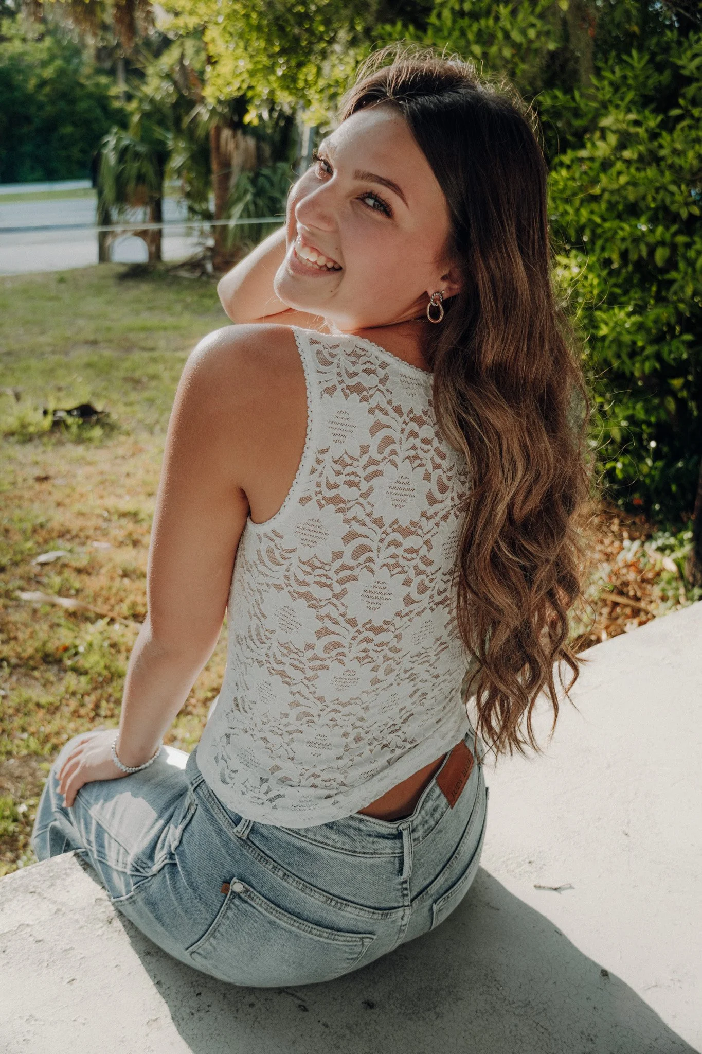 Young woman with long wavy brown hair, smiling and looking over her shoulder, wearing hoop earring, white lace sleeveless top, and blue jeans, sitting outdoors with green trees and shrubs in the background.