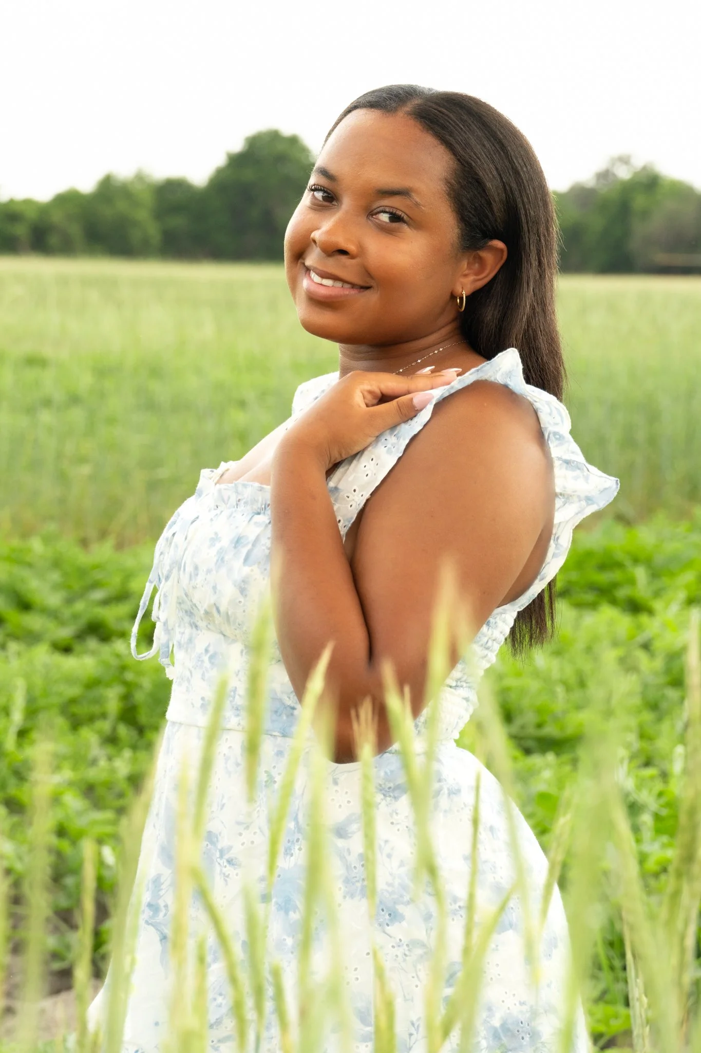 A young woman standing in a green field with her hand on her shoulder, smiling at the camera.