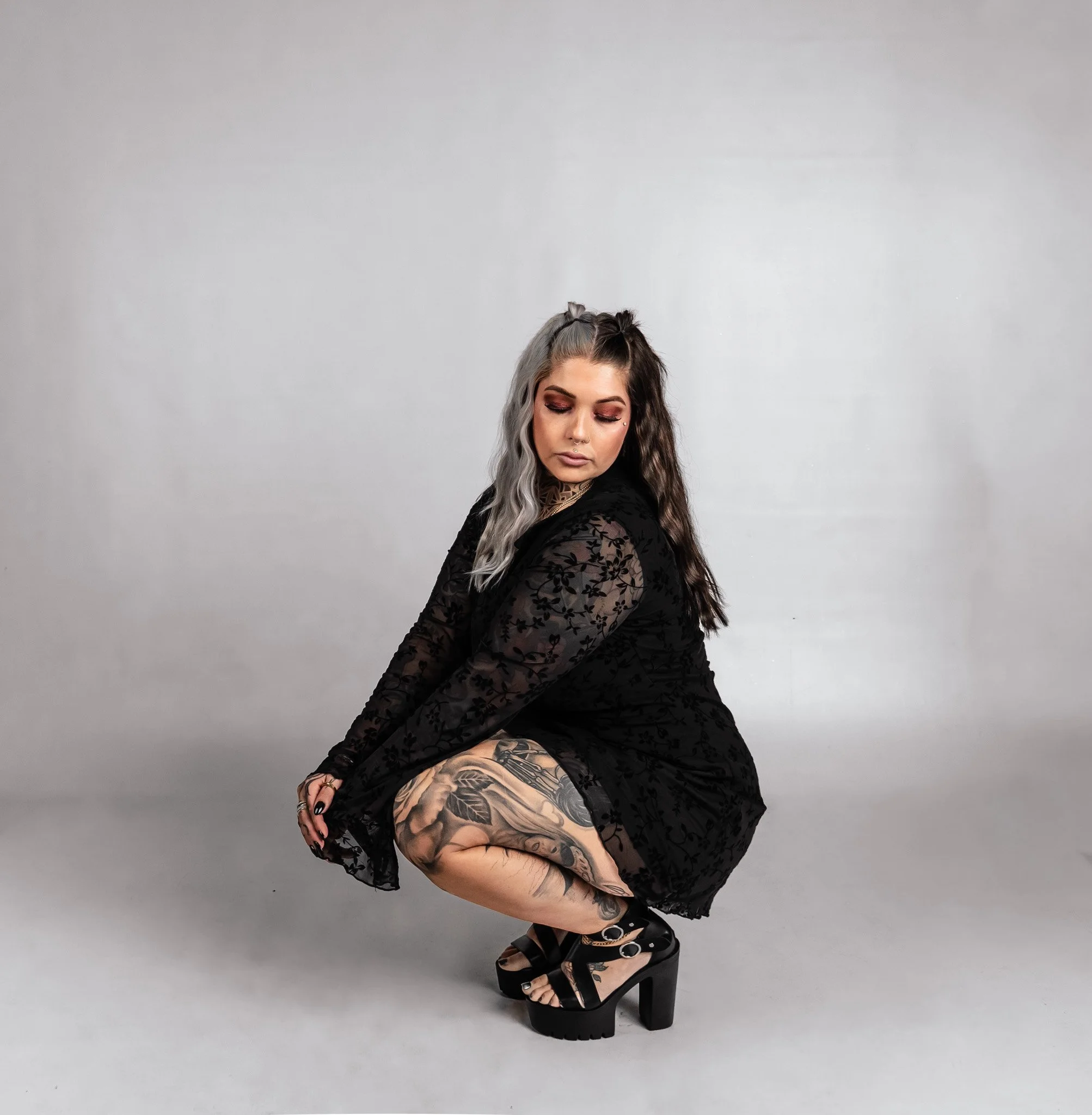 Woman crouching in a black lace dress, floral tattoos on her thigh, wearing black high-heeled sandals, with her eyes closed and hair half-gray, half-dark, against a plain gray background.