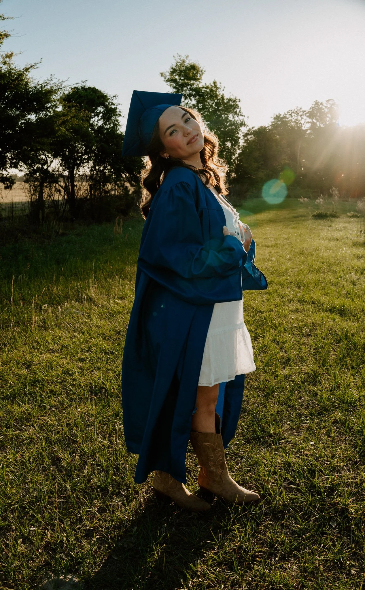 A woman in a blue graduation gown and cap standing in a grassy field with trees in the background, illuminated by sunlight.