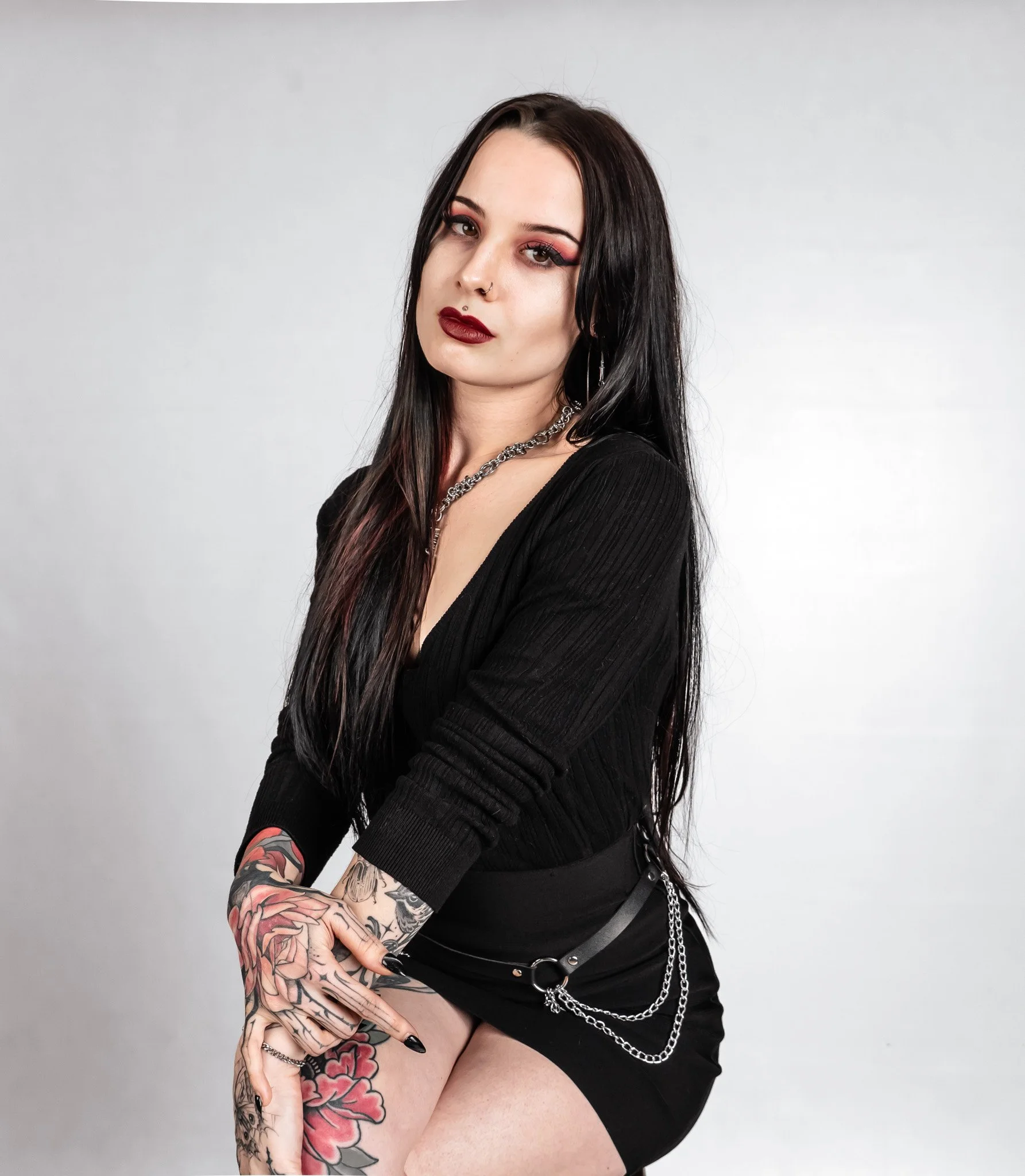A young woman with long black hair, tattoos on her arms and thigh, wearing dark makeup and a black top and skirt, posing against a plain background.