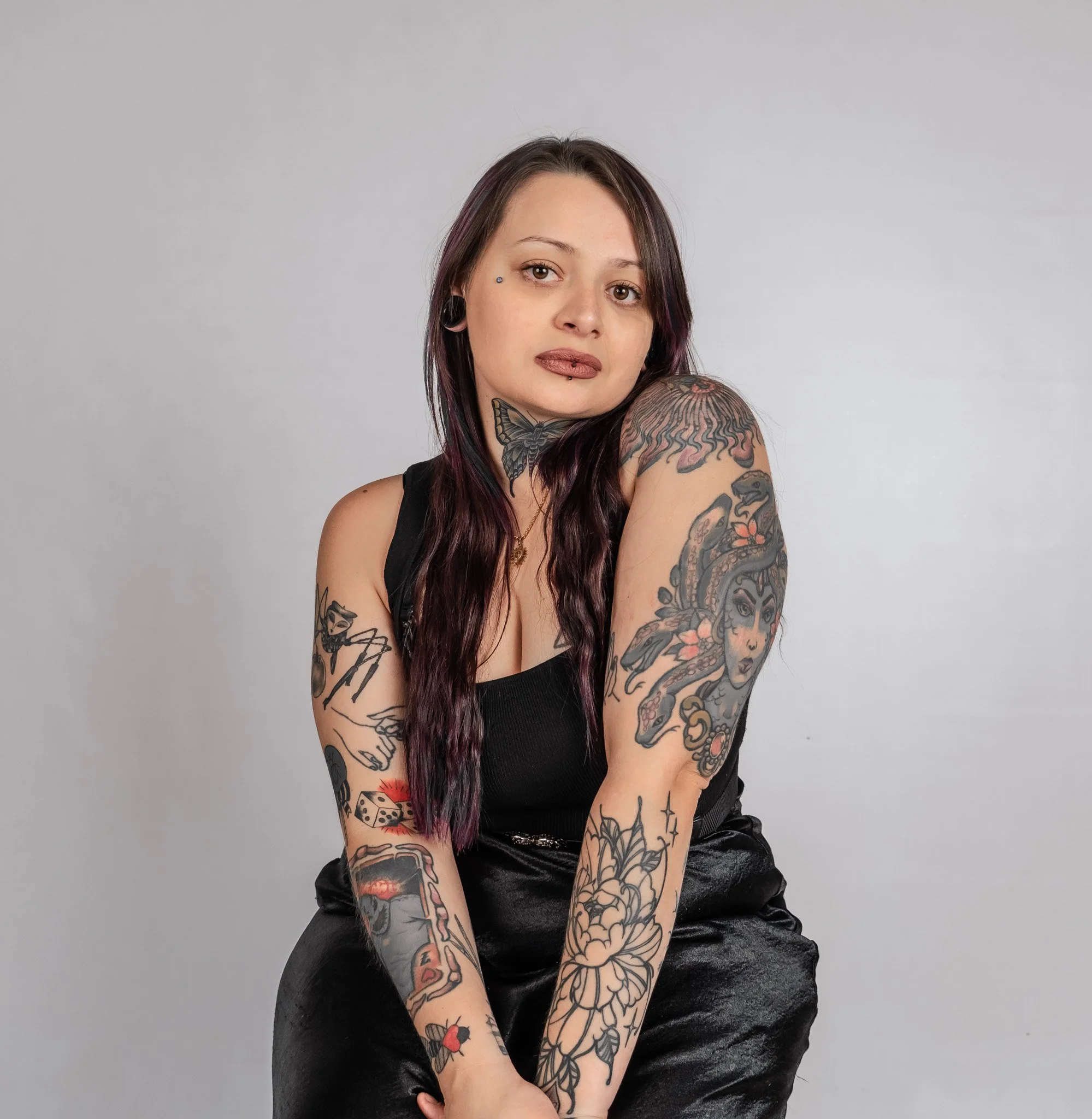 A young woman with long dark purple hair and tattoos on both arms and her chest, wearing a black top and black pants, looking at the camera against a plain gray background.