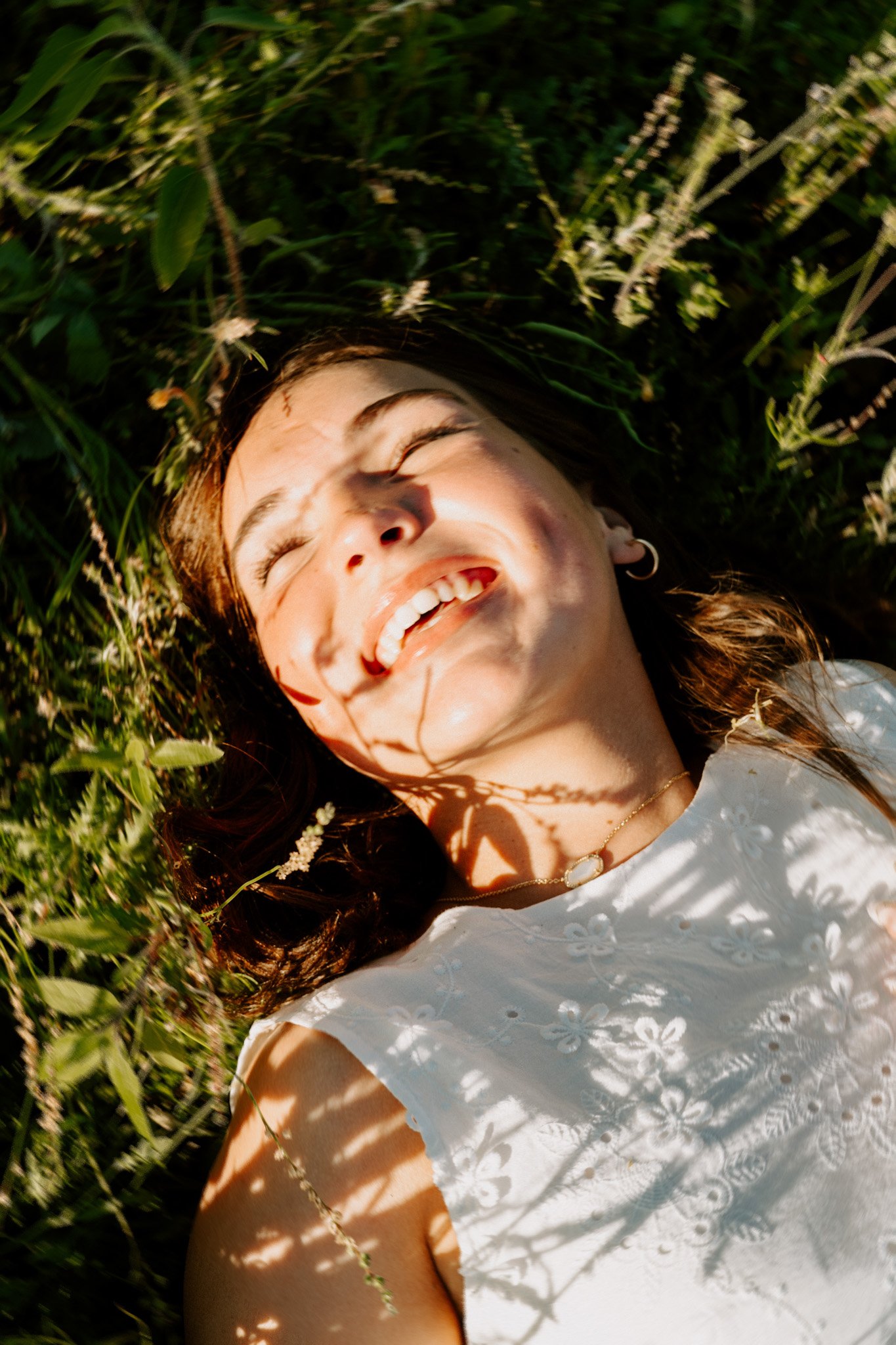 A woman with brown hair lying on green grass, smiling with her eyes closed, wearing a white embroidered dress and a necklace, with sunlight casting shadows around her.