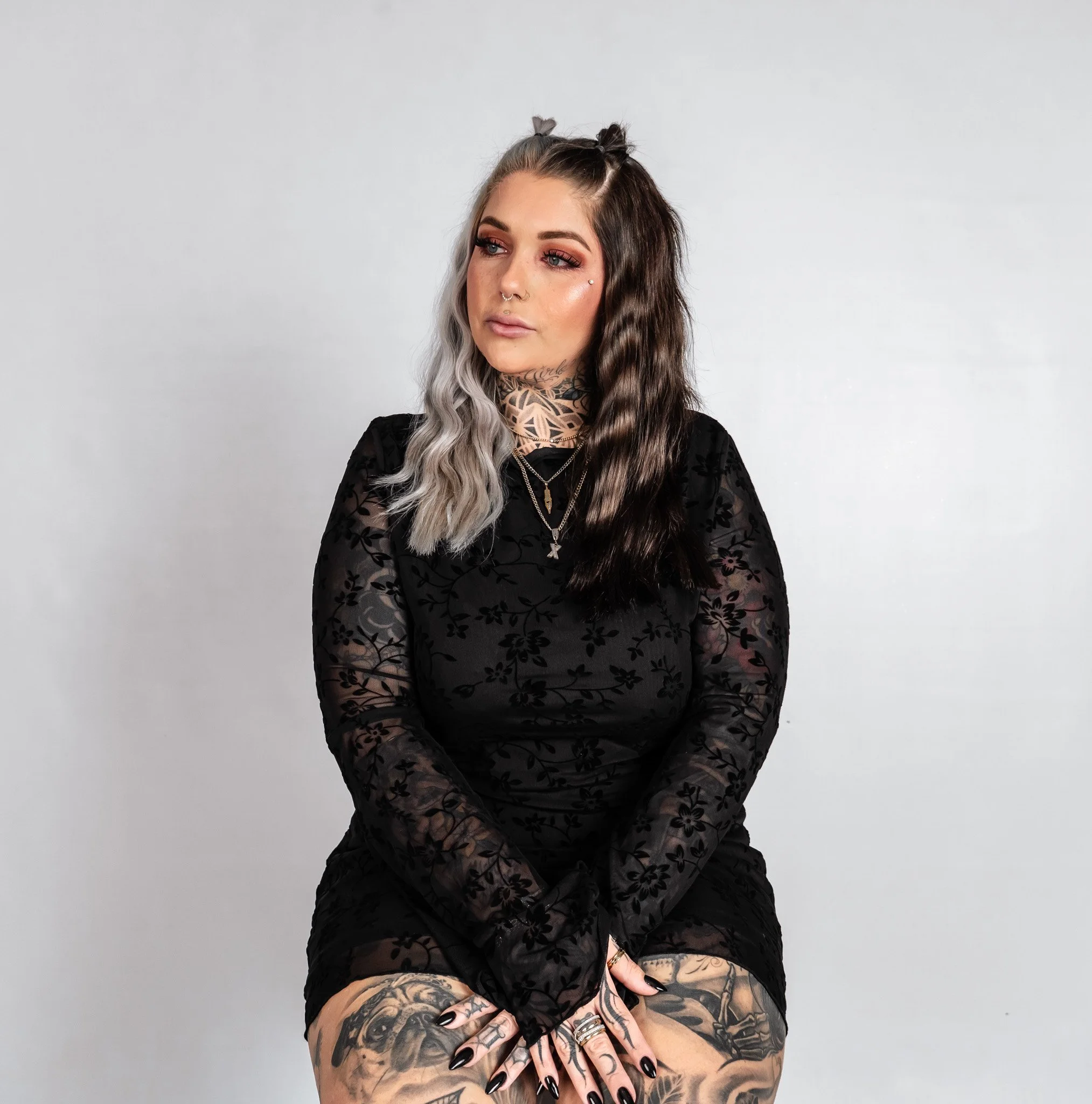 A young woman with mixed blonde and dark hair, tattoos, and jewelry, wearing a black floral dress, sitting against a plain white background.