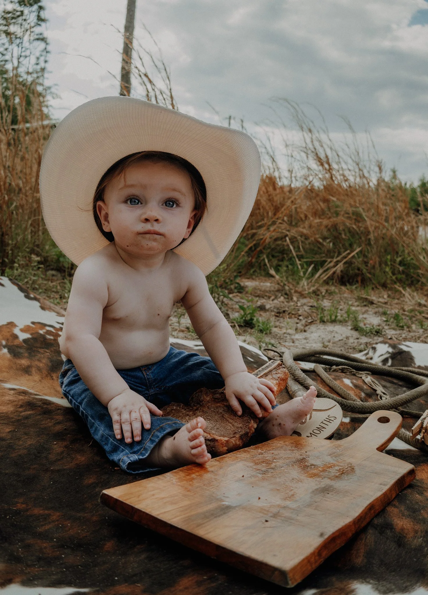 A young boy with a large sunhat sits on a/log on a rustic wooden table outdoors, surrounded by natural landscape and cloudy sky.