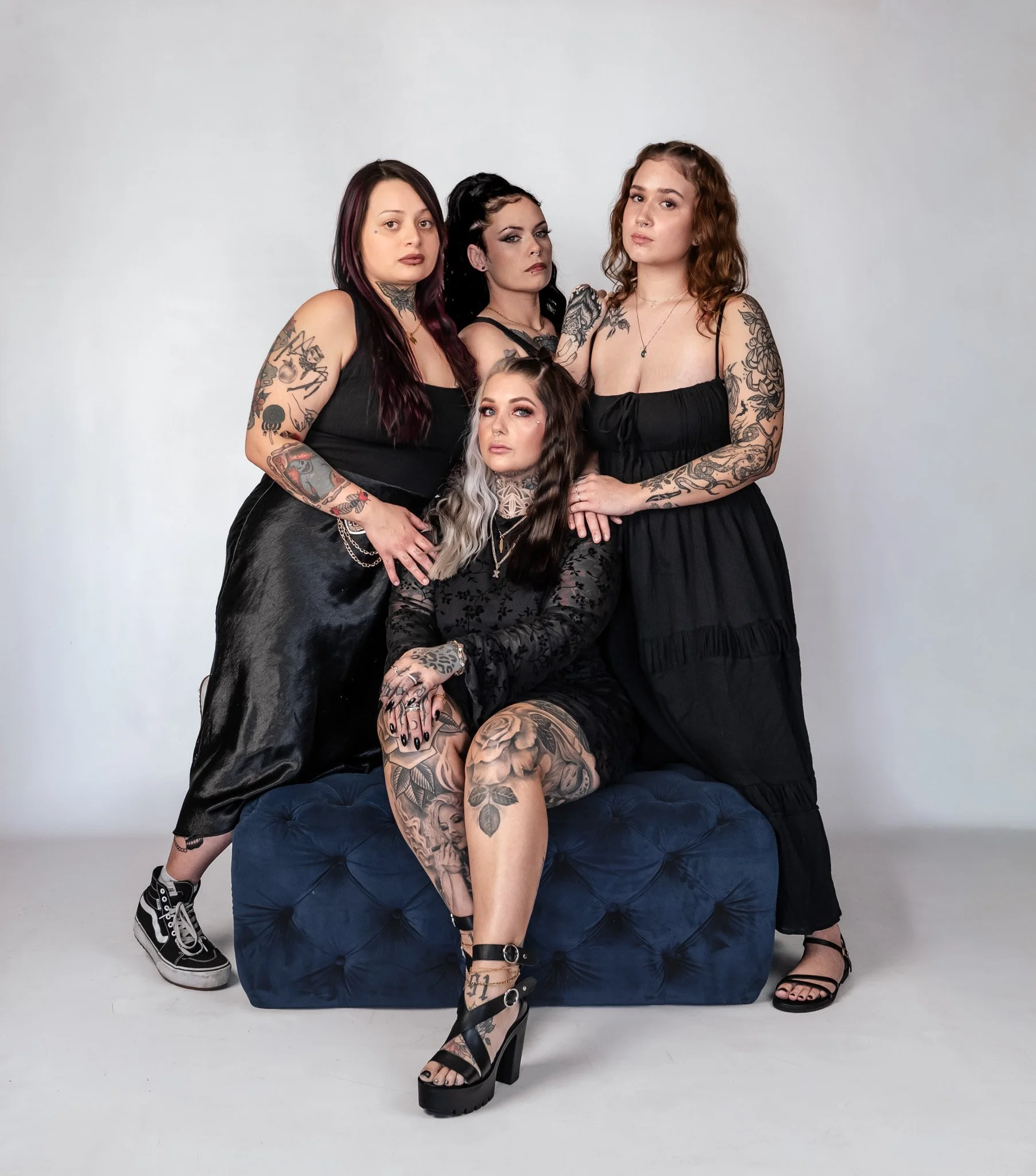Four women with tattoos in black dresses posing on a blue velvet couch, with a plain white background.