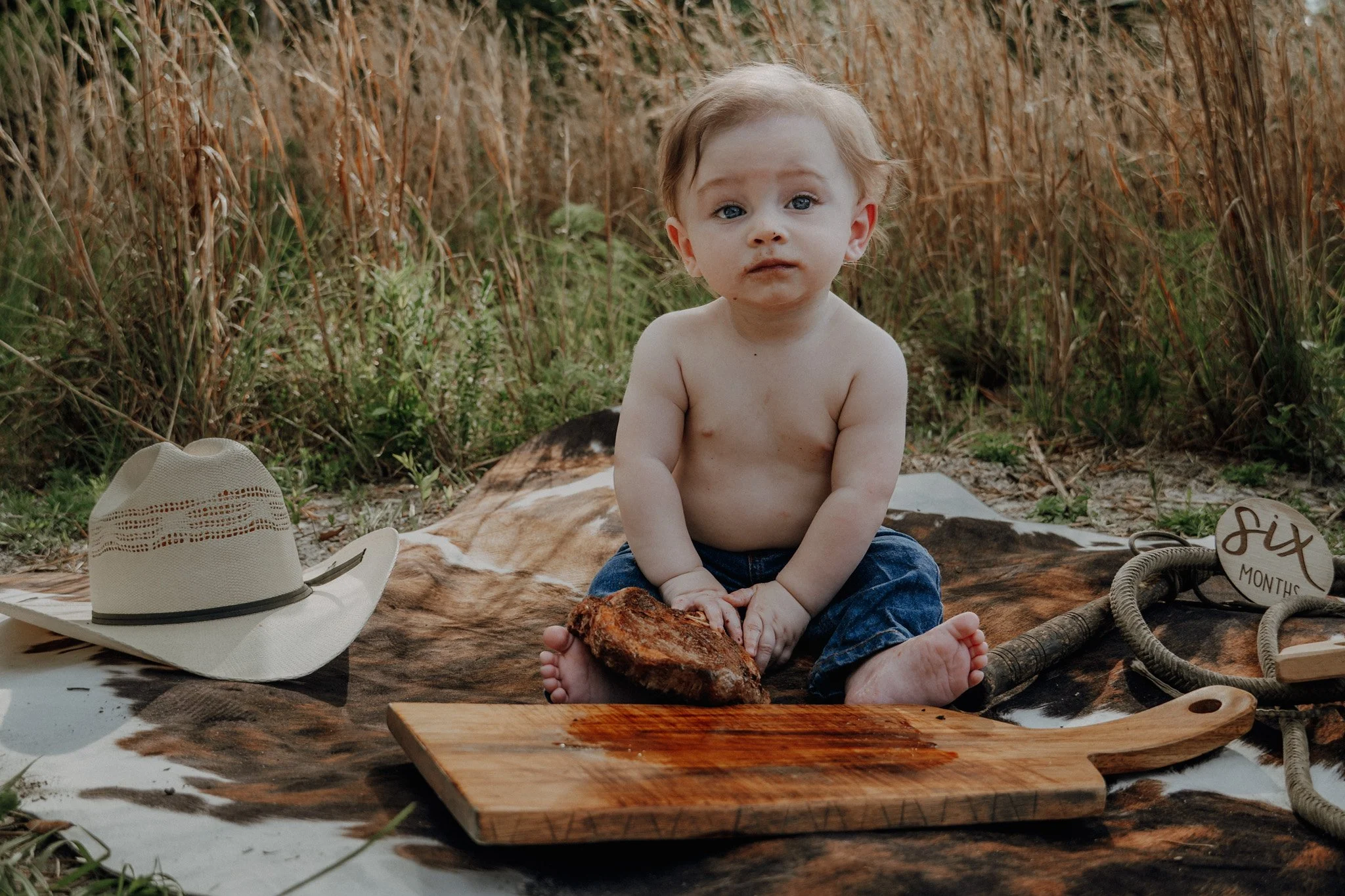 A young shirtless boy with light hair and blue eyes sitting on a blanket outdoors amid dried tall grass with accessories around, including a white cowboy hat, a small round sign reading 'six months,' and a wooden cutting board with a piece of meat, p
