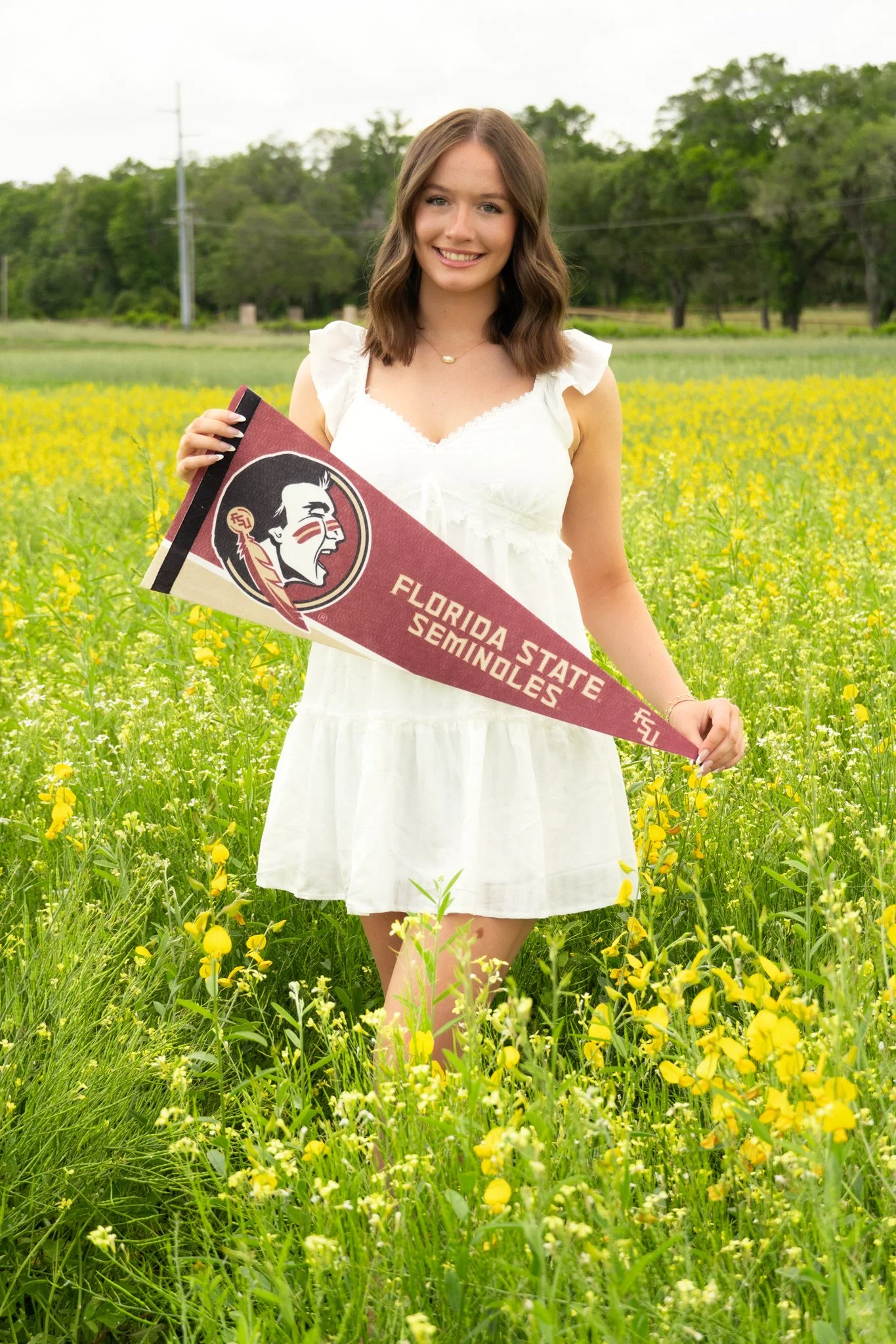 Young woman standing in a field of yellow flowers, holding a Florida State Seminoles pennant, smiling, wearing a white dress.
