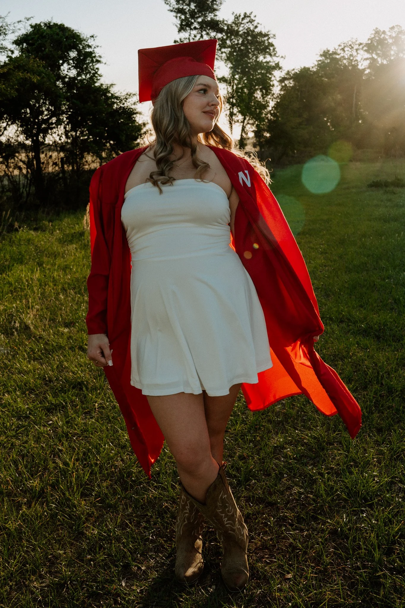 A young woman in a white dress and cowboy boots stands in a grassy field at sunset, wearing a red graduation gown and cap. She looks to the side with a smile, with trees and the sun in the background.