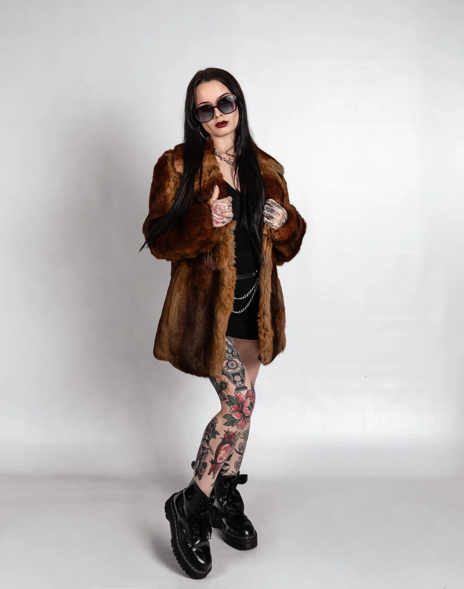 Woman with long dark hair wearing sunglasses, a fur coat, black dress, combat boots, and has tattoos on her legs and hands, standing against a plain white background.