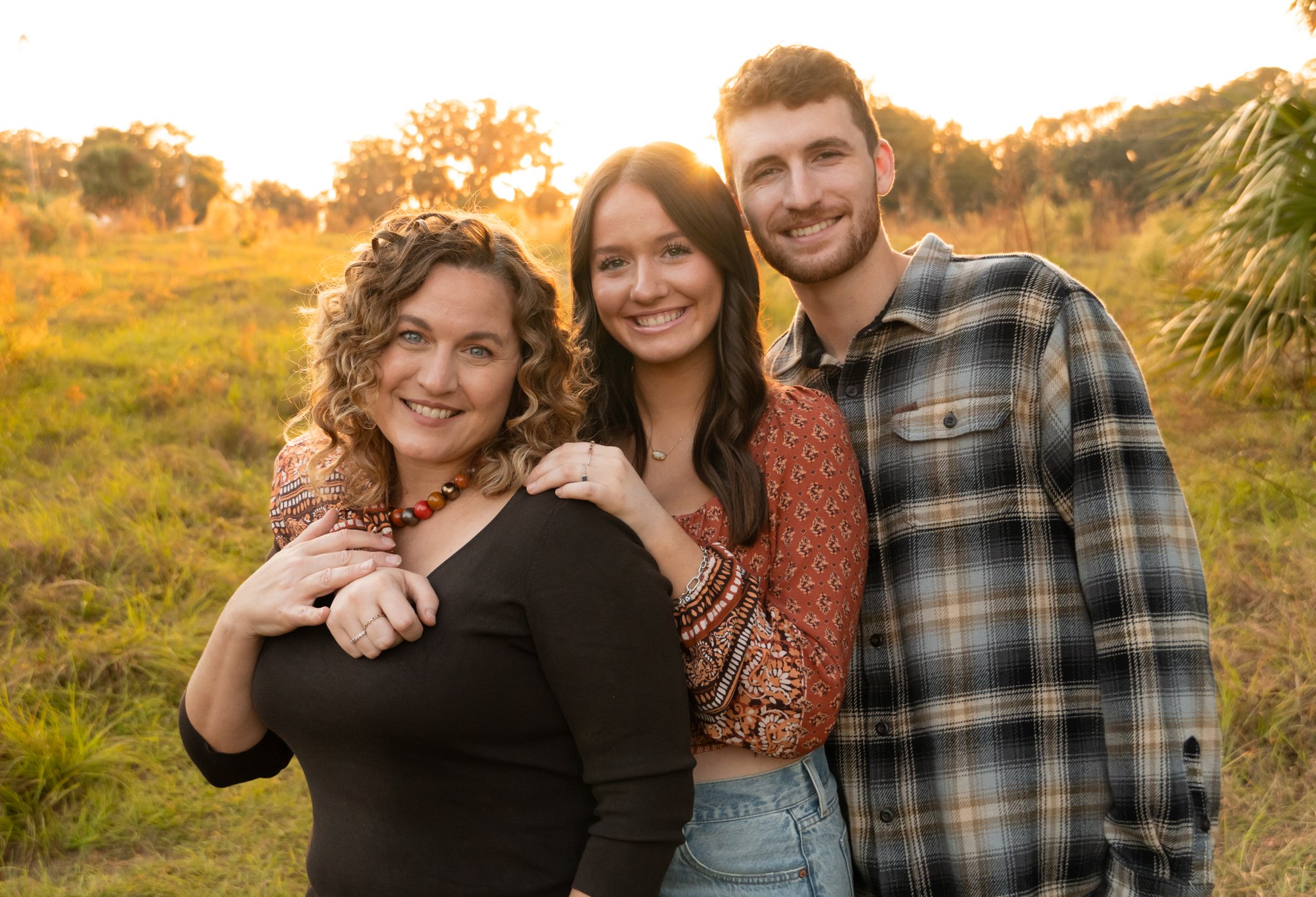 A group of three young adults smiling, close together outdoors during sunset, with autumn trees and grass in the background.