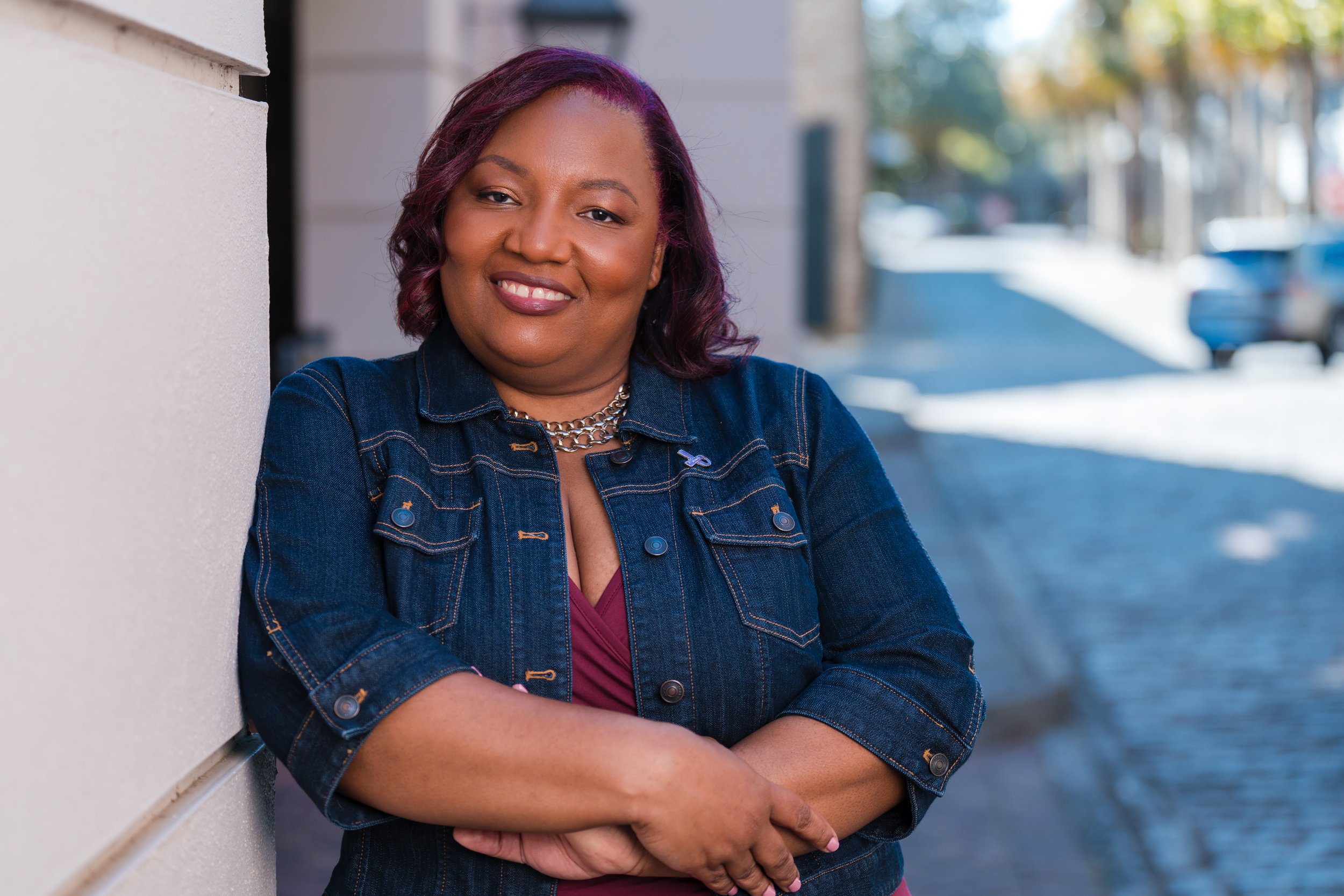 A woman with brown hair and dark skin leaning against a wall outdoors, smiling, wearing a denim jacket and a maroon top with a chain necklace, with a blurred street scene in the background.