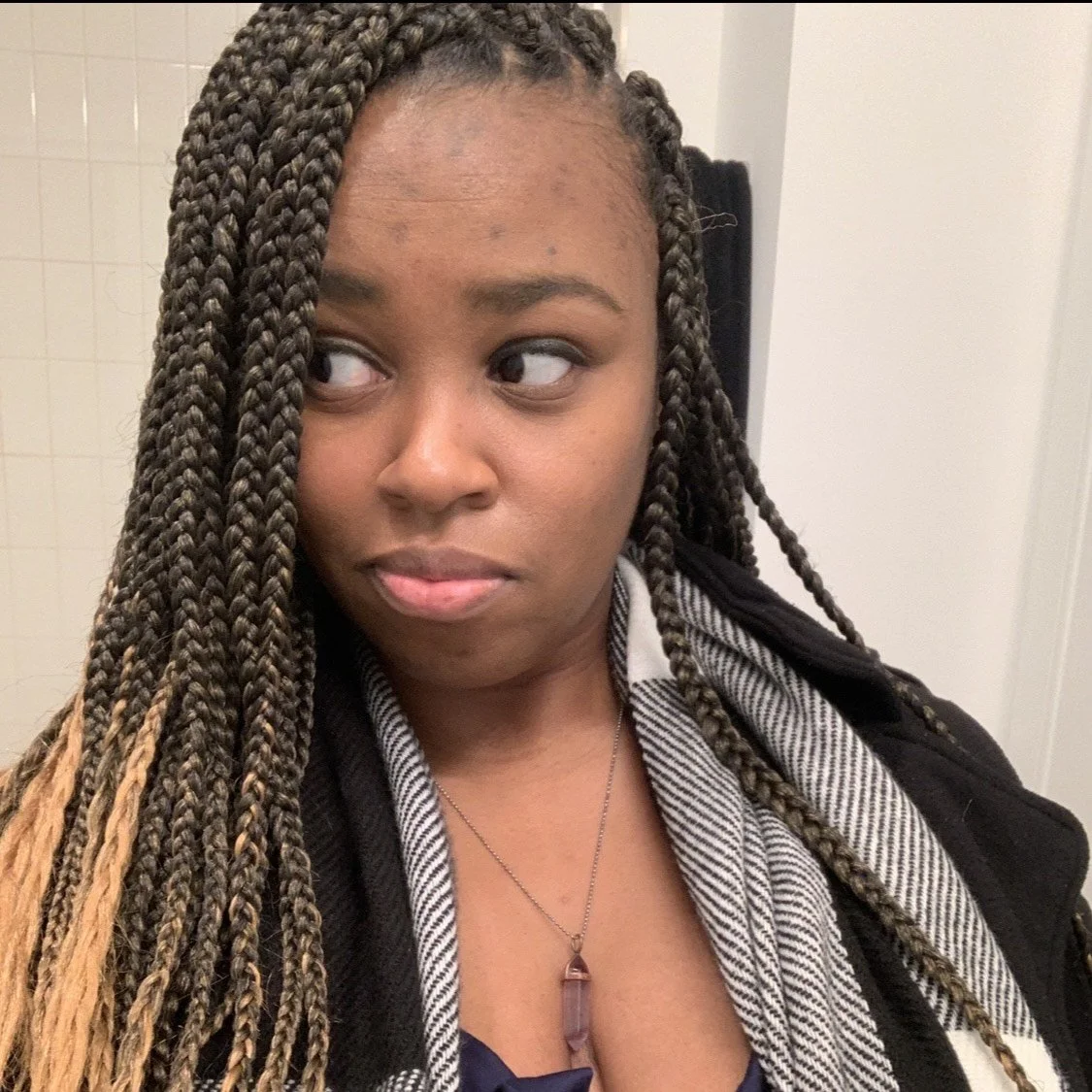 A woman with long braided hair looking to her left, wearing a striped shirt, a black jacket, and a pendant necklace, in an indoor setting.