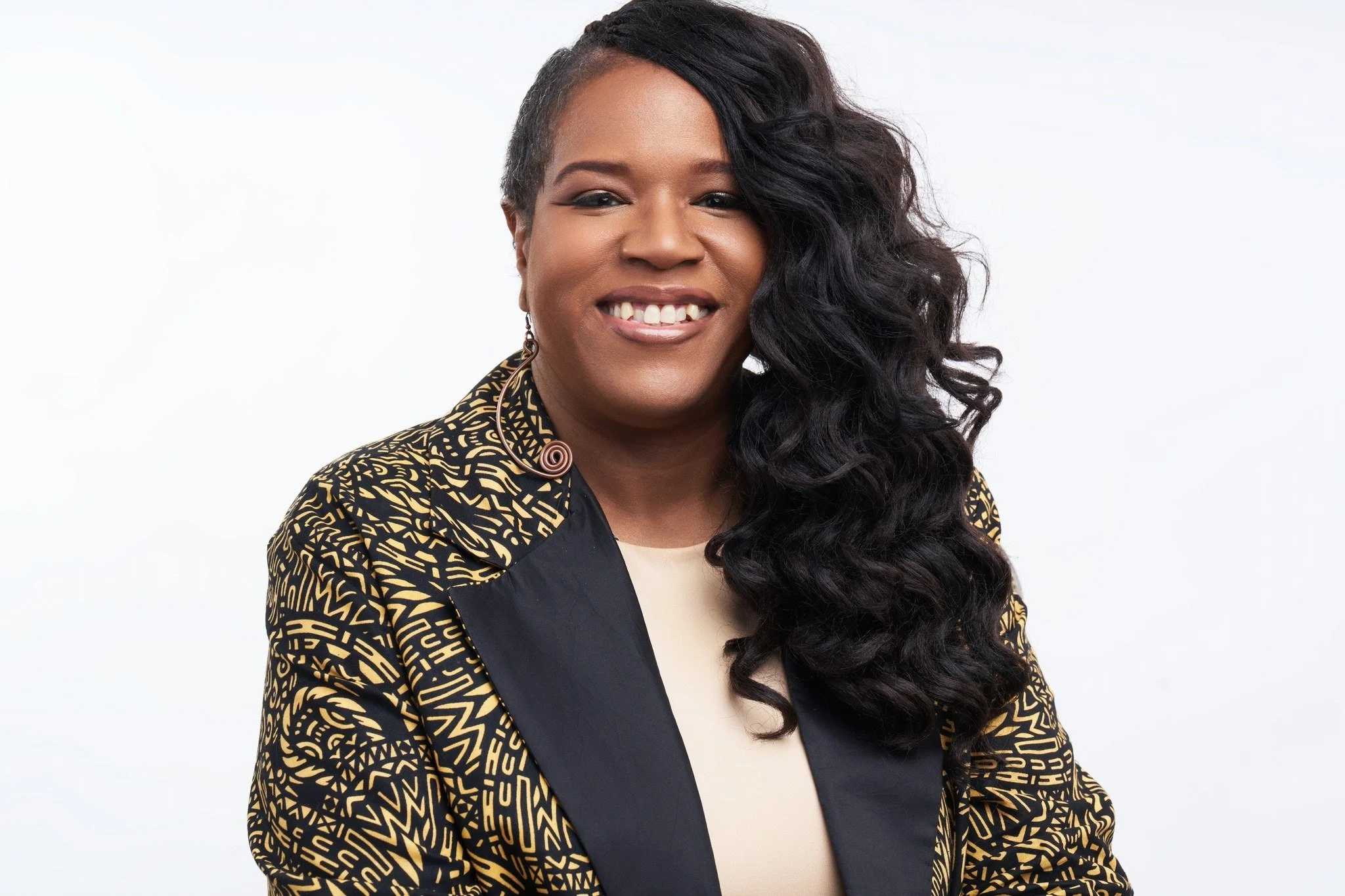 Portrait of a smiling woman with long curly black hair, wearing a patterned blazer and earrings, against a plain white background.