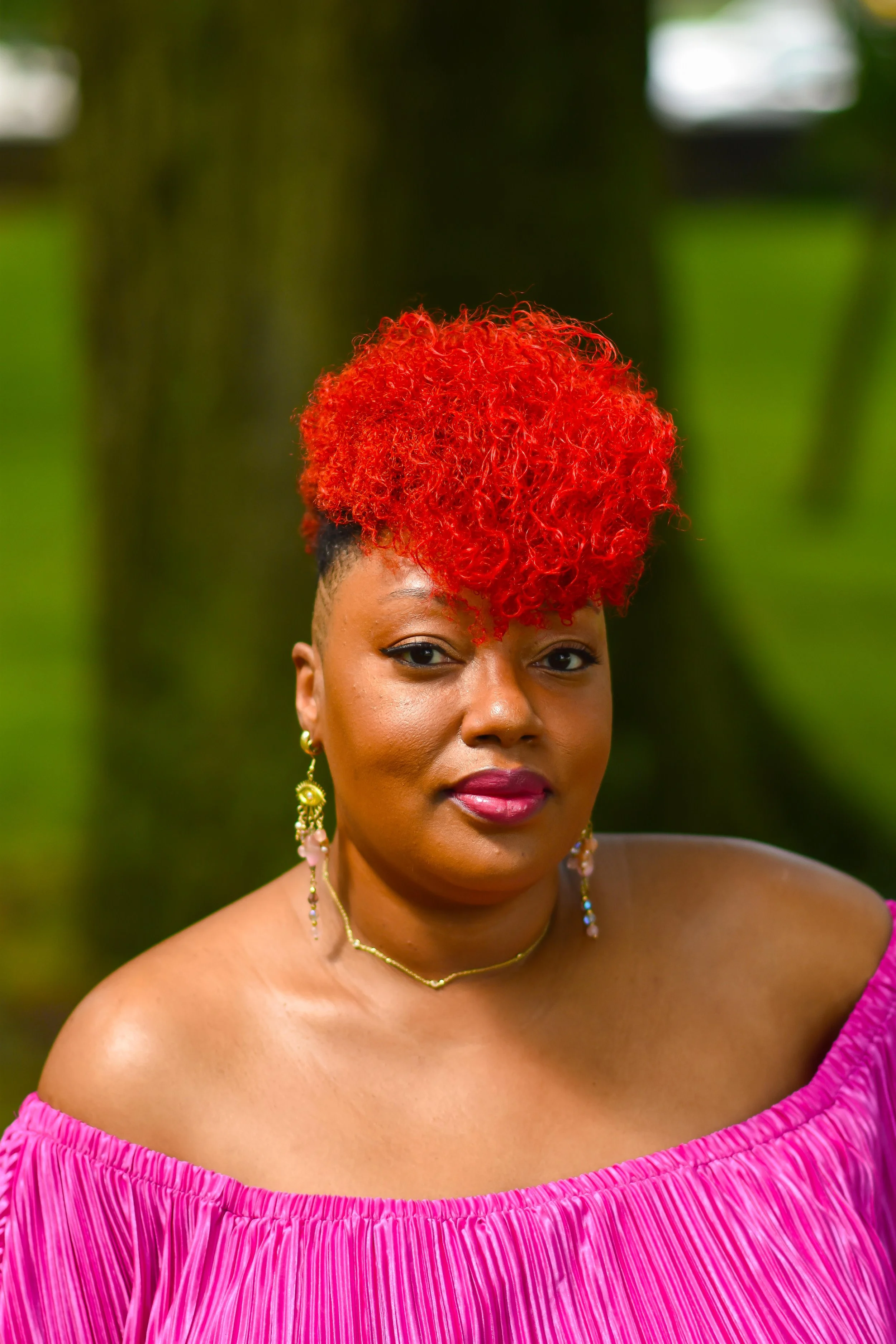 A woman with long earrings and a gold necklace, wearing an off-the-shoulder pink dress, with red curly hair, standing outdoors with trees in the background.