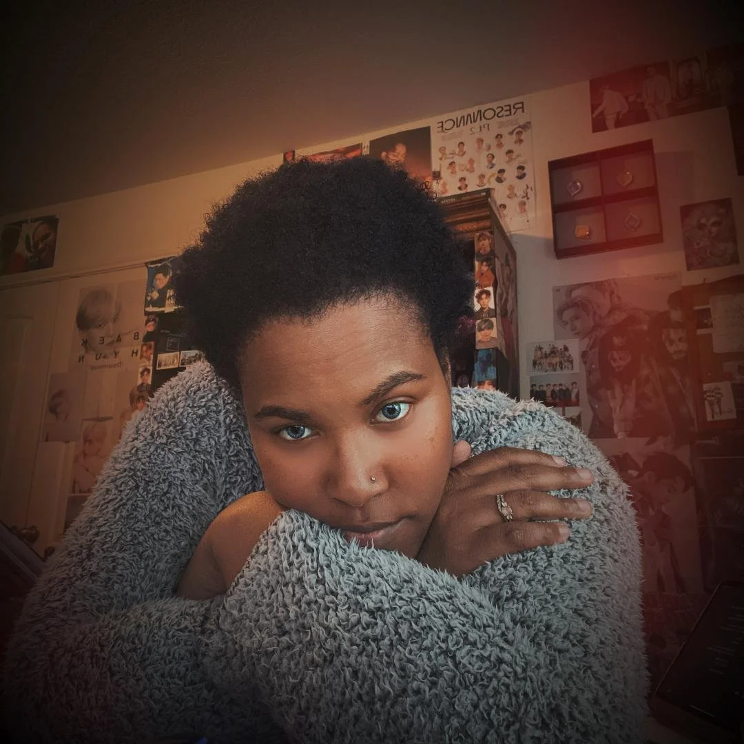 A woman with short curly hair and blue eyes resting her chin on crossed arms wrapped in a gray, fluffy sweater, looking directly at the camera in a room decorated with posters and pictures.
