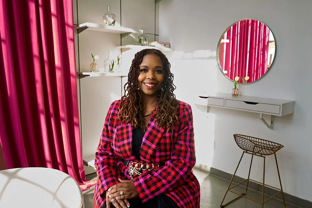 A woman with long curly hair and a bright smile, wearing a red and black plaid blazer, sitting in a modern room with white walls and pink curtains.