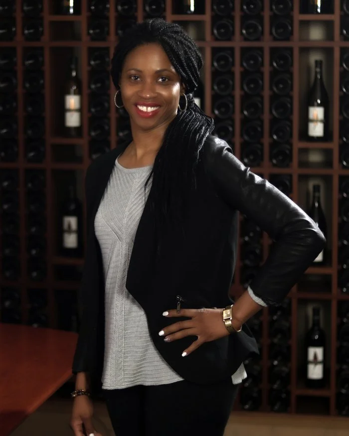 A woman with braided hair is standing in front of a wine rack, smiling, wearing a black leather jacket, a light gray sweater, and gold jewelry.