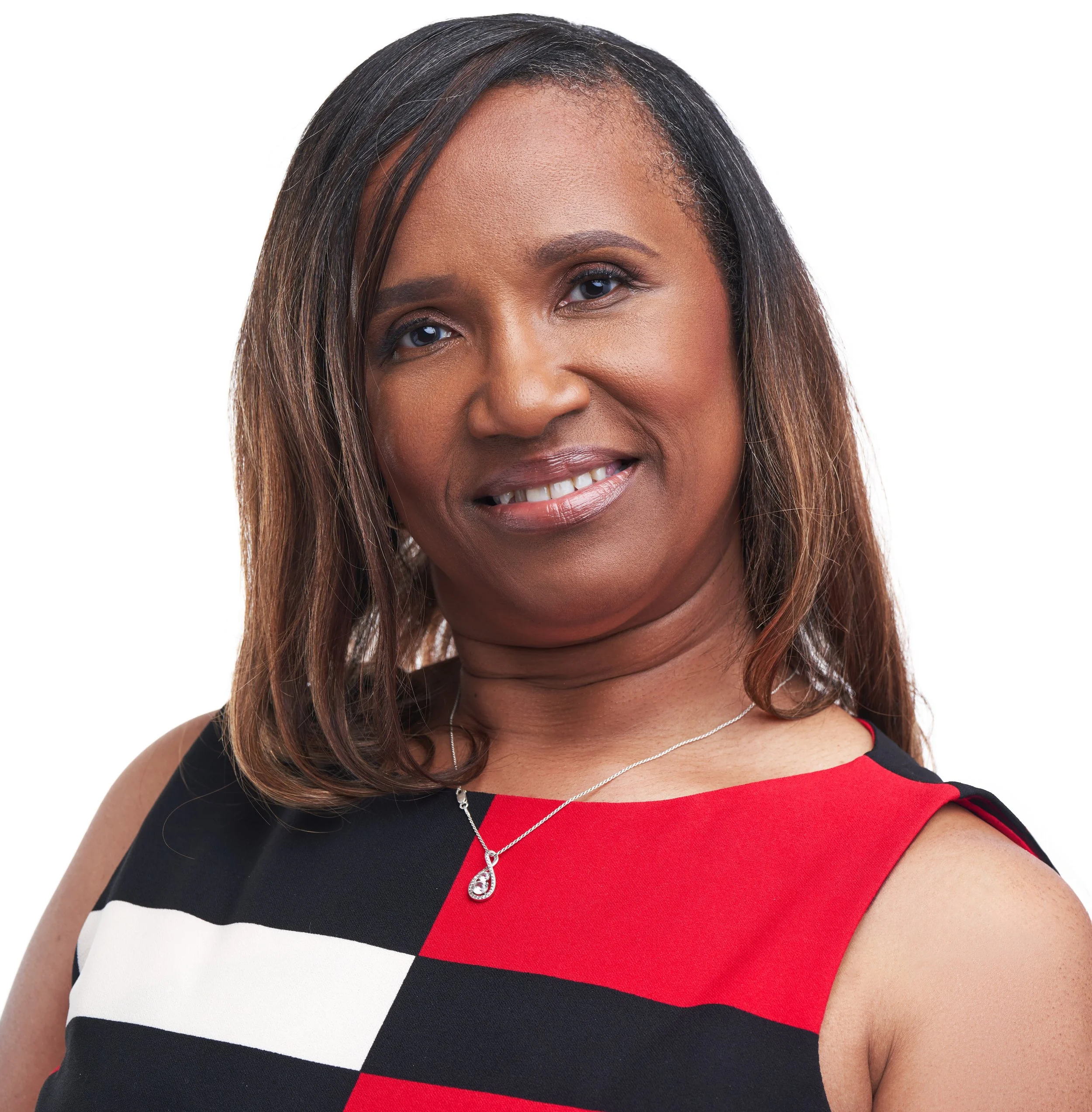 A smiling woman with medium skin tone, shoulder-length straight hair, wearing a sleeveless dress with a black, white, and red color block pattern, and a silver necklace with a teardrop pendant.