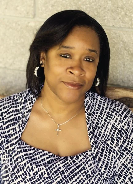 A woman with shoulder-length black hair, wearing hoop earrings, a cross necklace, and a patterned top, sitting against a grey brick wall background.