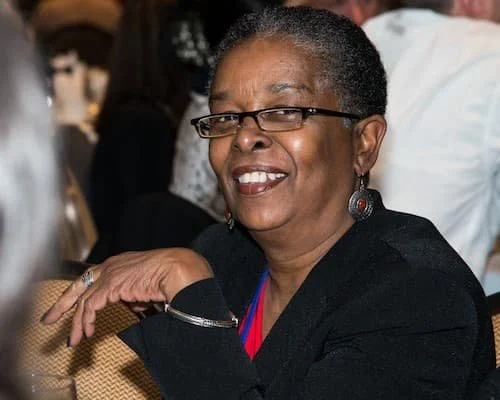 Smiling woman with short hair, glasses, and earrings, sitting at a table in what appears to be a social gathering or event.