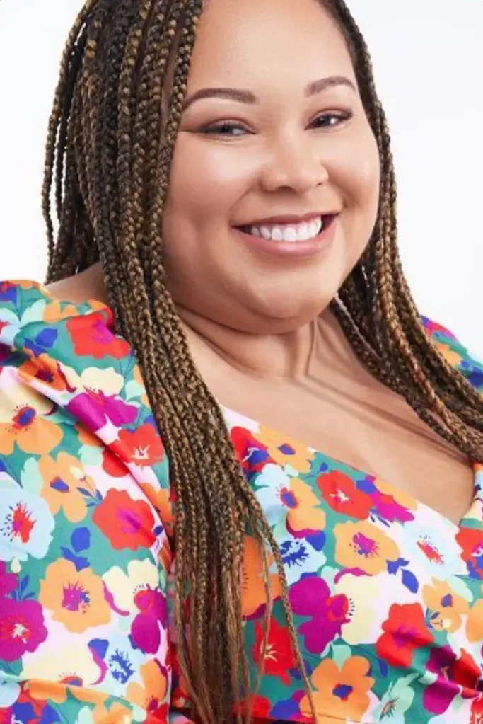 Close-up of a smiling woman with braided hair and earrings, indoor setting with a window in the background.