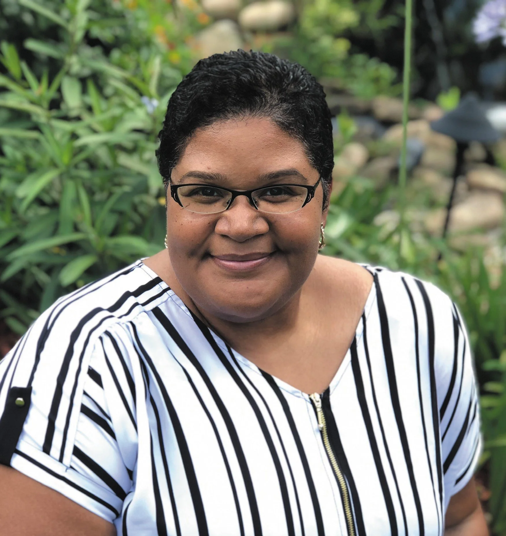 A smiling woman with short curly black hair, wearing glasses and a white shirt with black vertical stripes, standing outdoors in front of green plants.