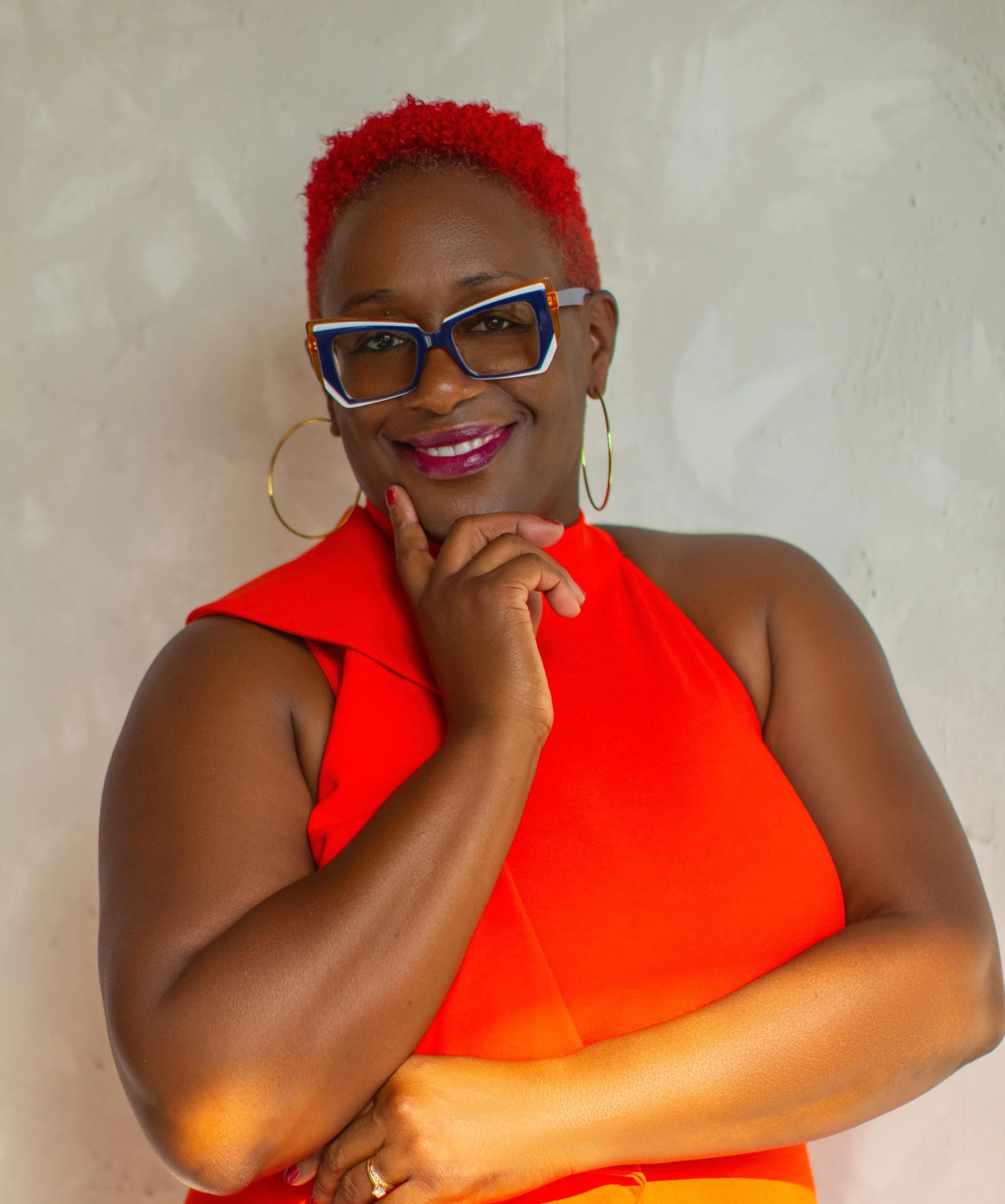 A woman with short, curly red hair wearing large blue glasses, gold hoop earrings, a red sleeveless top, and lipstick, smiling and posing with her hand on her chin against a neutral background.