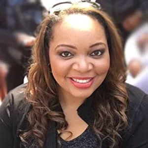 Close-up of a smiling woman with wavy brown hair and light makeup, wearing a black top and a black blazer.