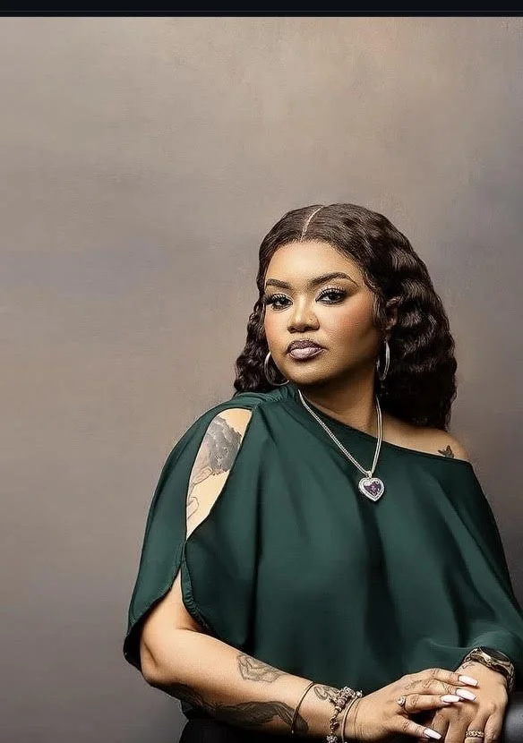 Portrait of a woman with dark wavy hair, wearing a dark green top with cut-out shoulders, jewelry including a heart-shaped pendant necklace, hoop earrings, and bracelets, standing against a plain background.