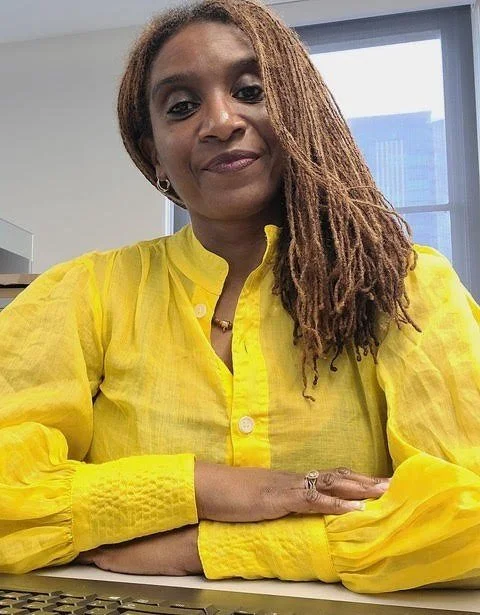 A woman with long, light brown dreadlocks, wearing a yellow blouse with long sleeves, sitting at a desk in an office with large windows and city buildings in the background.