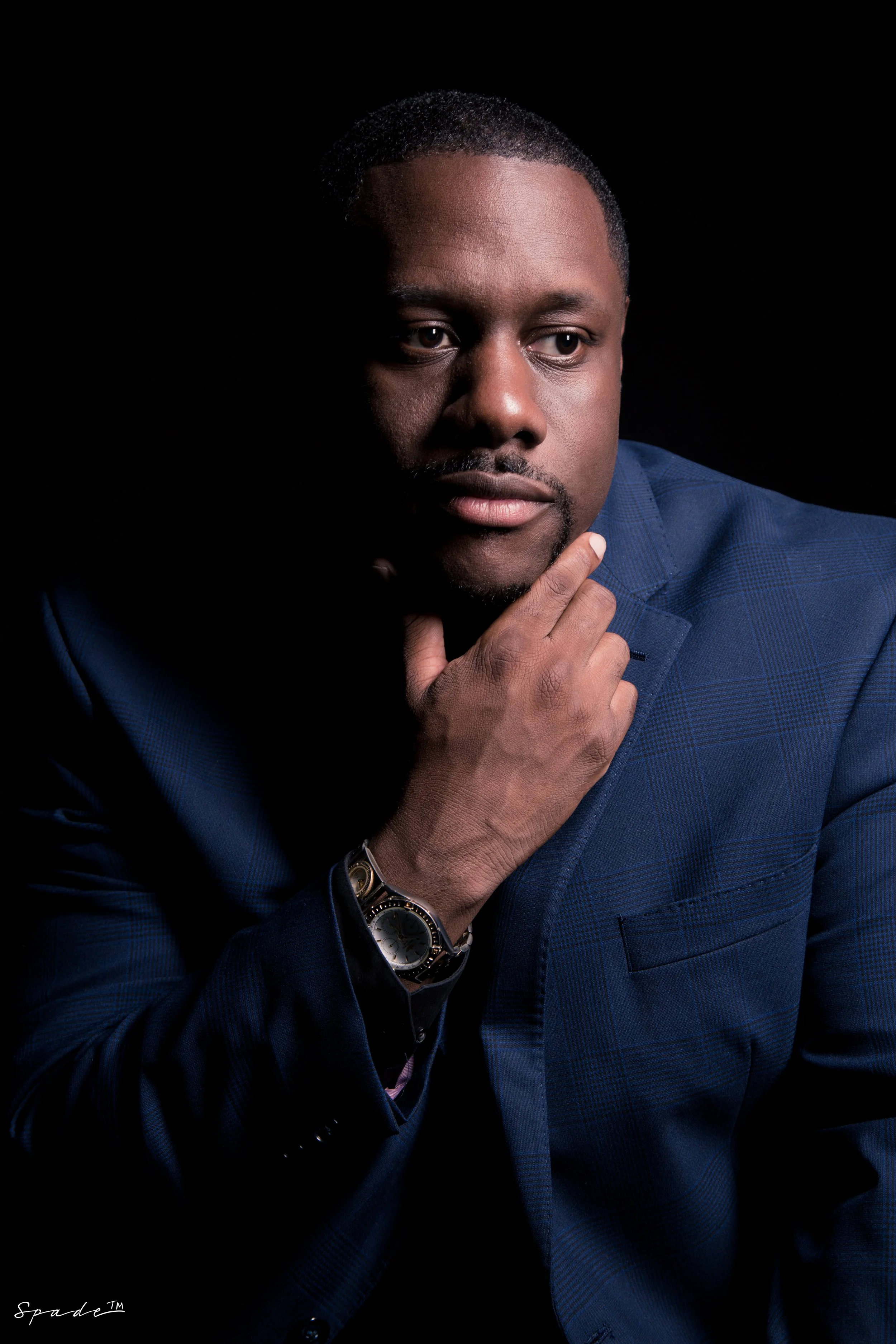 A man in a blue suit ponders, resting his chin on his hand, against a black background.
