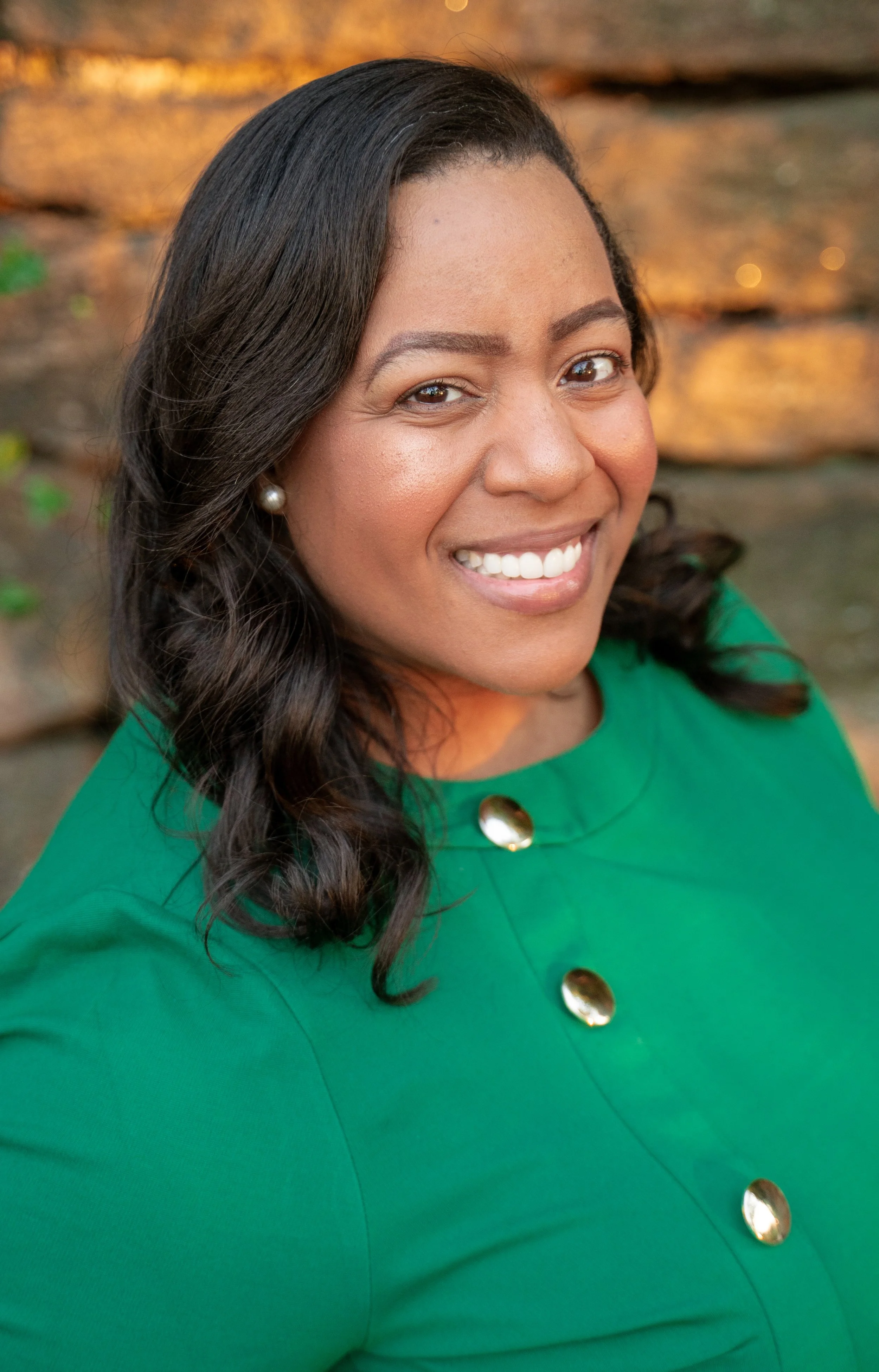 Smiling woman with dark wavy hair, wearing a green top with gold buttons, standing outdoors with a blurred wood background.