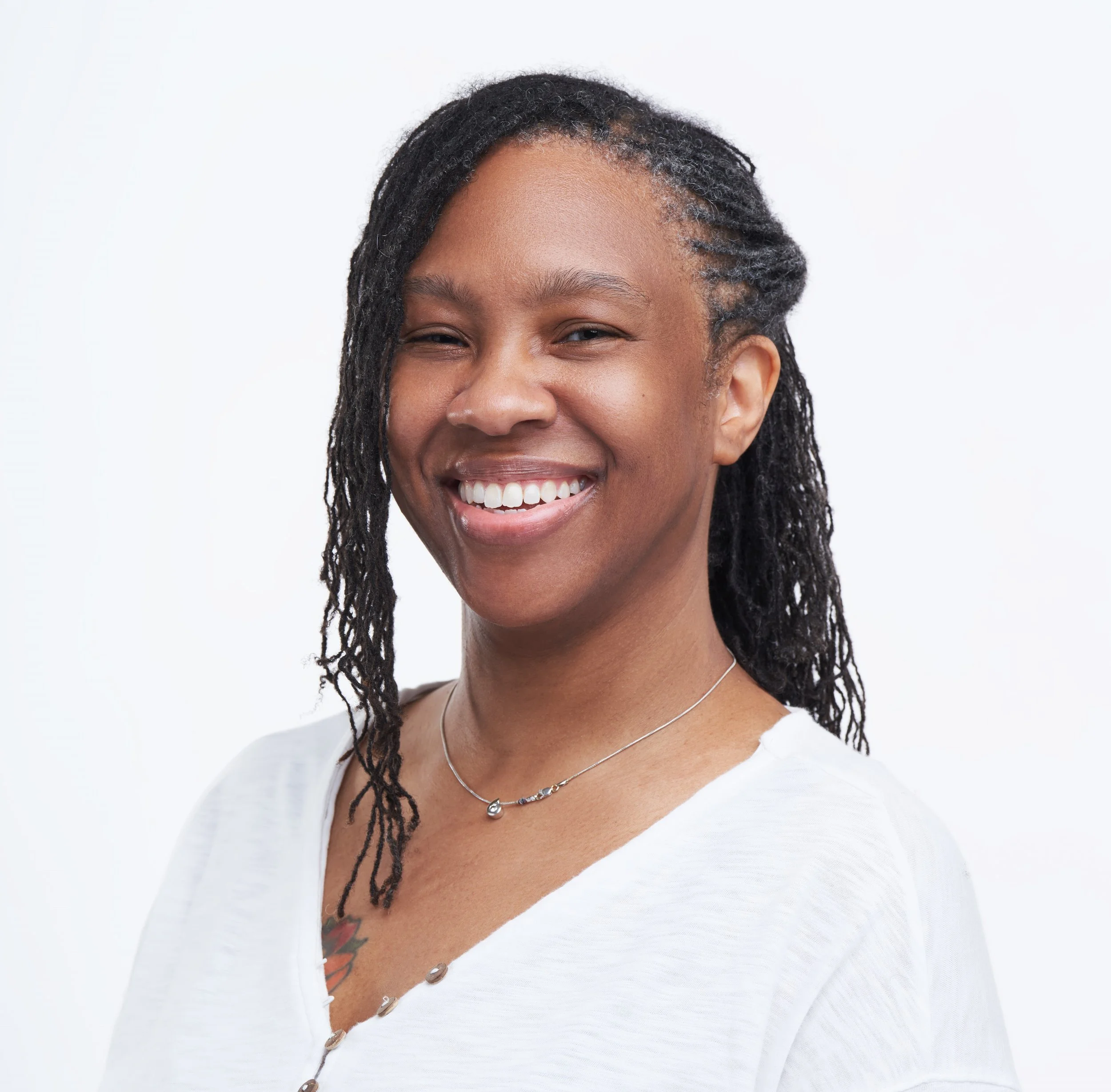 Portrait of a smiling woman with dreadlocks wearing a white top and a delicate necklace, against a plain white background.