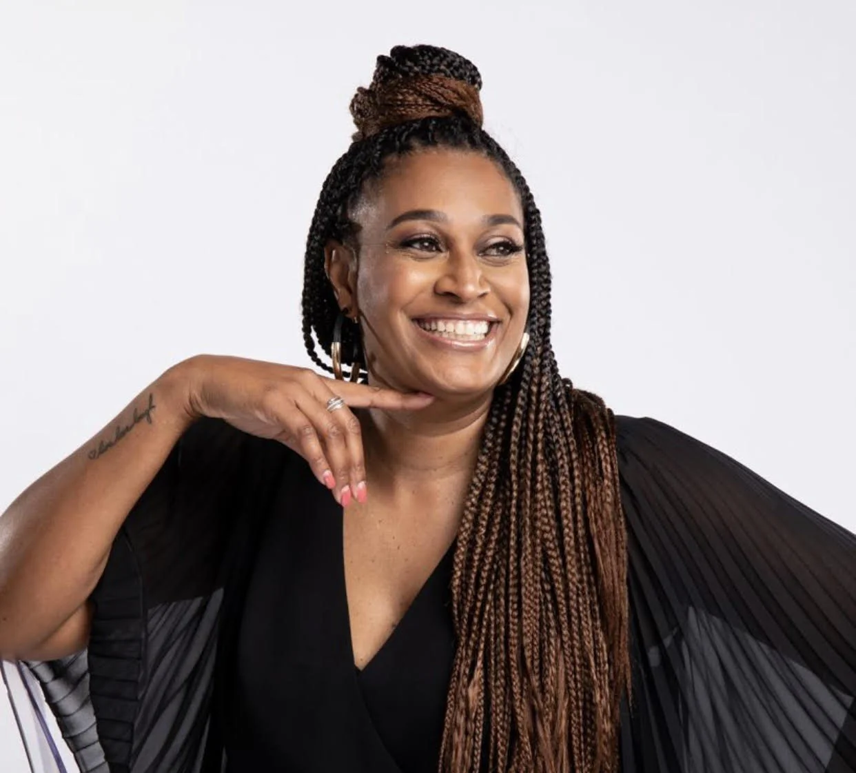 A woman with braided hair in a bun smiling and posing with her hand near her chin against a plain white background.