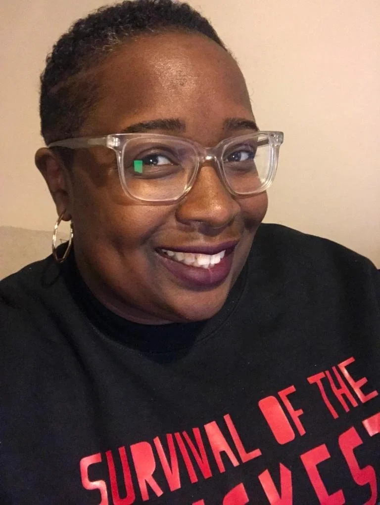 A smiling person with short curly hair, wearing glasses and a black shirt with red text, indoors against a plain wall.