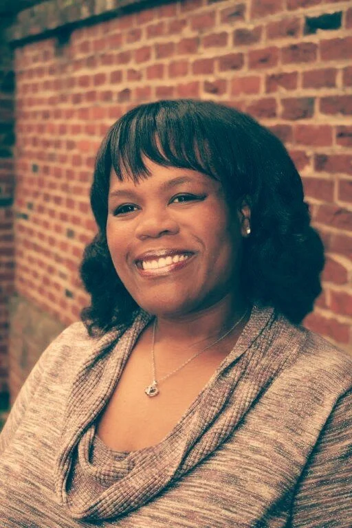 Smiling woman with black hair and pearl earrings, wearing a gray top, in front of a brick wall.