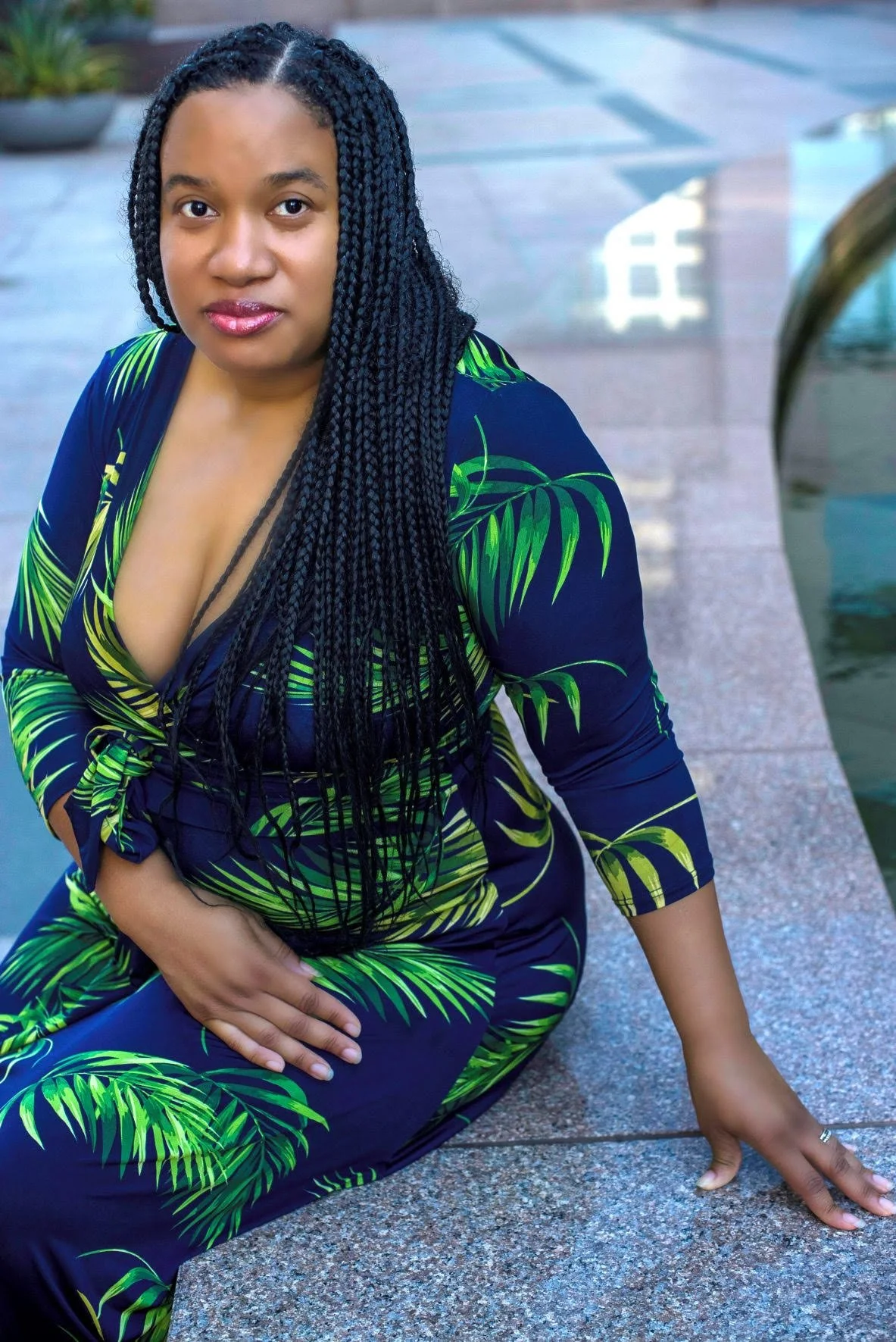 A woman with long braided hair sitting by a water feature outside, wearing a navy blue dress with green tropical leaf patterns, posing with one hand on her knee and the other on the ground, looking directly at the camera.