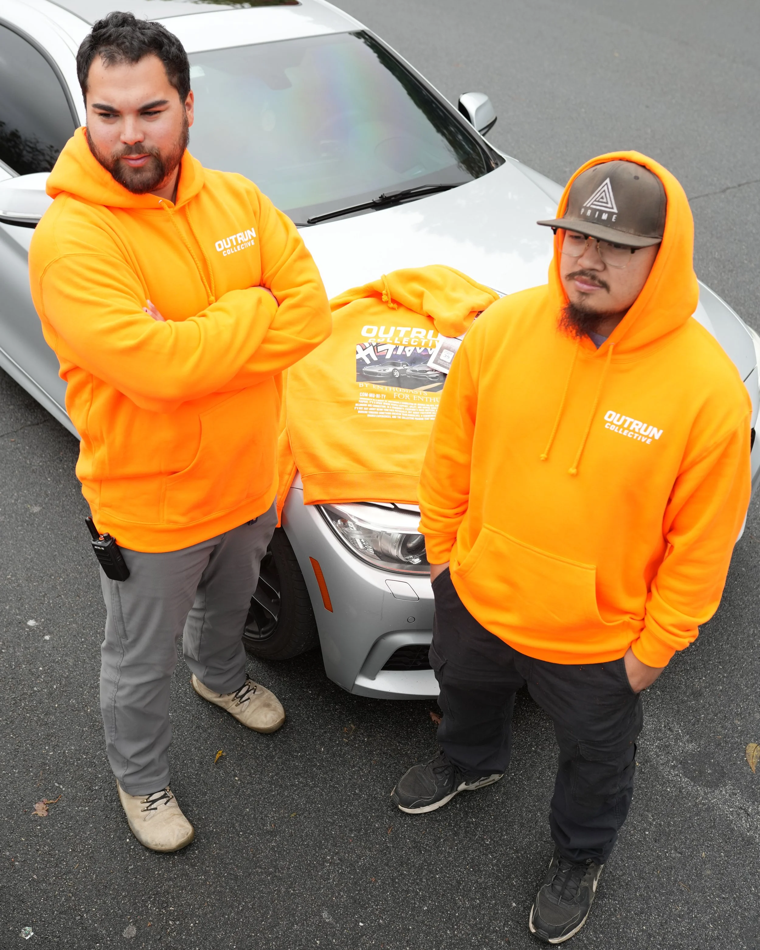 Two men standing beside a silver car, both wearing bright orange hoodies with 'OUTRUN COLLECTIVE' printed on them. One man has crossed arms, the other has hands in pockets. An orange hoodie is spread out on the hood of the car, and a t-shirt with similar design is on top of it.