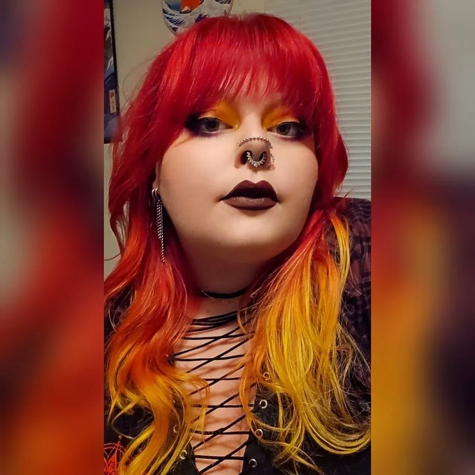 Person with red and yellow hair, dark lipstick, and bold eye makeup, wearing nose and ear piercings, and a black lace-up top.