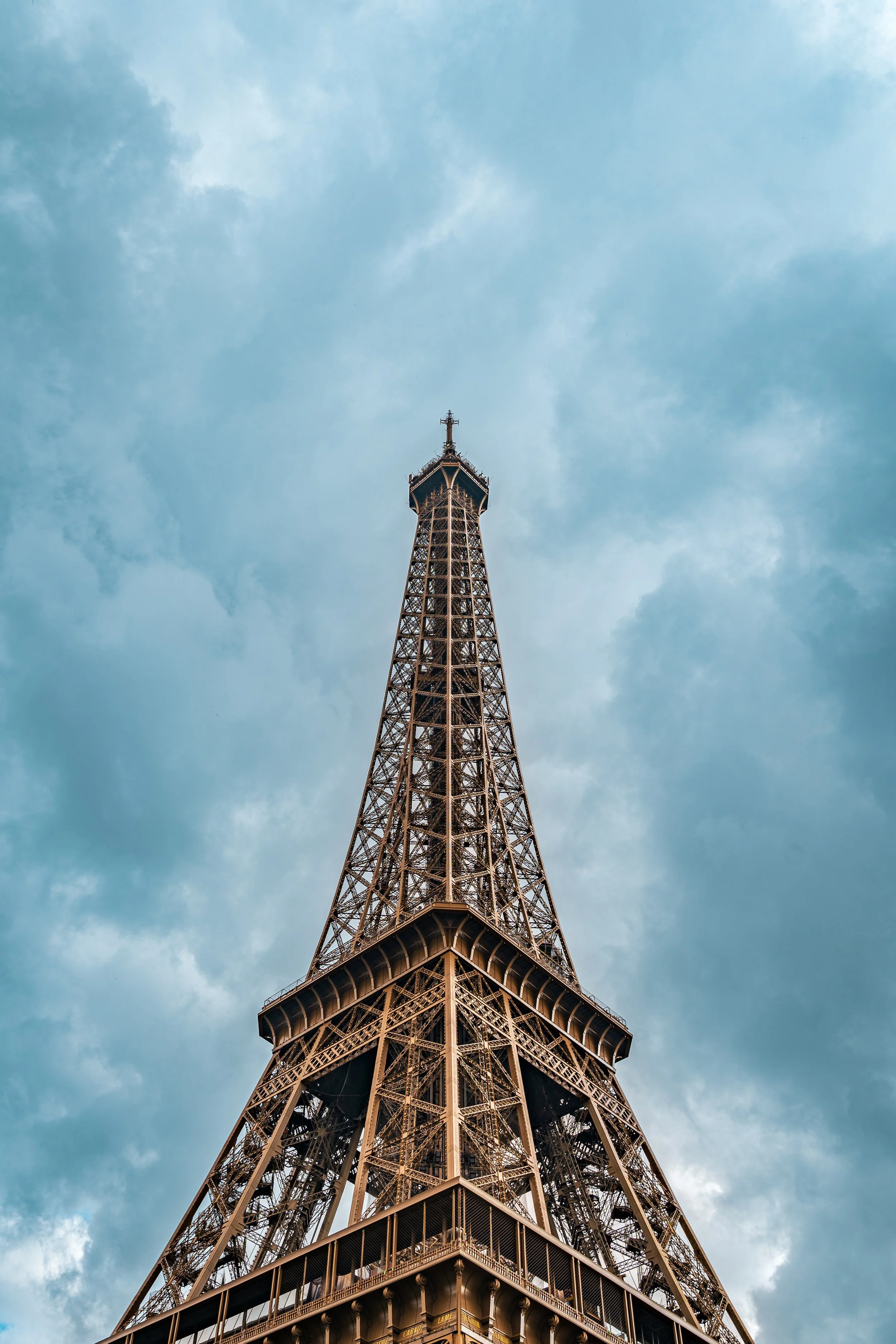 Looking up toward the Eiffel Tour in Paris, which is placed in the center of the frame. The blue sky contrasts with the copper metalwork of this iconic Parisian landmark. Paris, France, 2023.