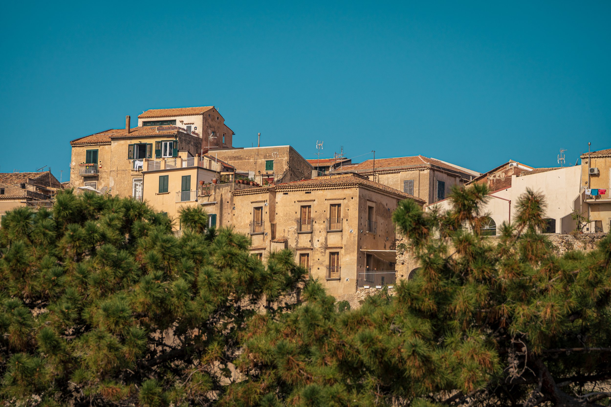 Coppery-brown buildings on the cliffs of Tropea, Italy. Out-of-focus trees are in the foreground. The sky is a bright blue in the background.